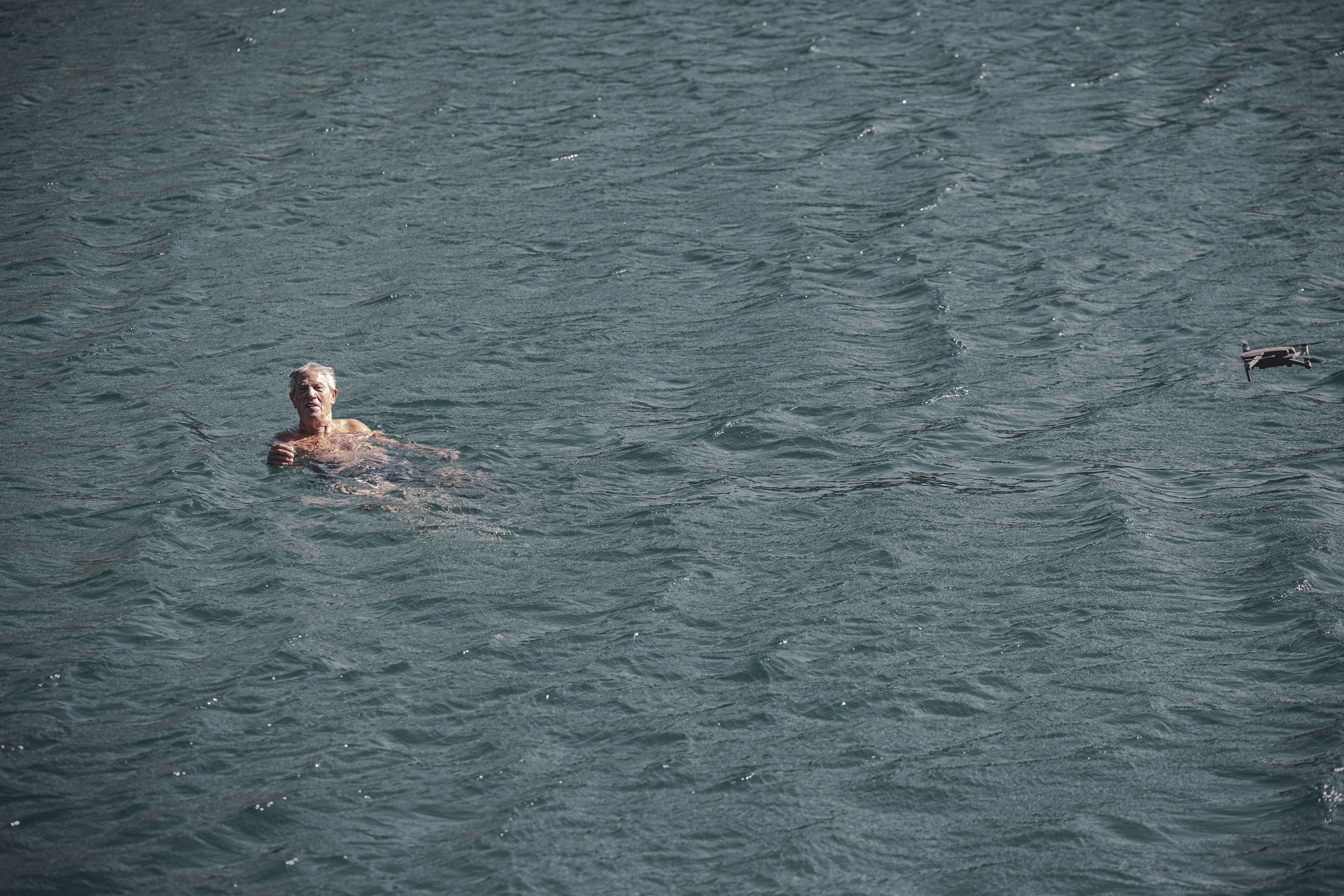Eine Person schwimmt in einem Gewässer mit einem dunkelblauen Farbton, begleitet von einem kleinen, schwimmenden Vogel in der Nähe rechts. Das Wasser ist leicht gekräuselt und reflektiert Licht. © Fotografie Tomas Rodriguez