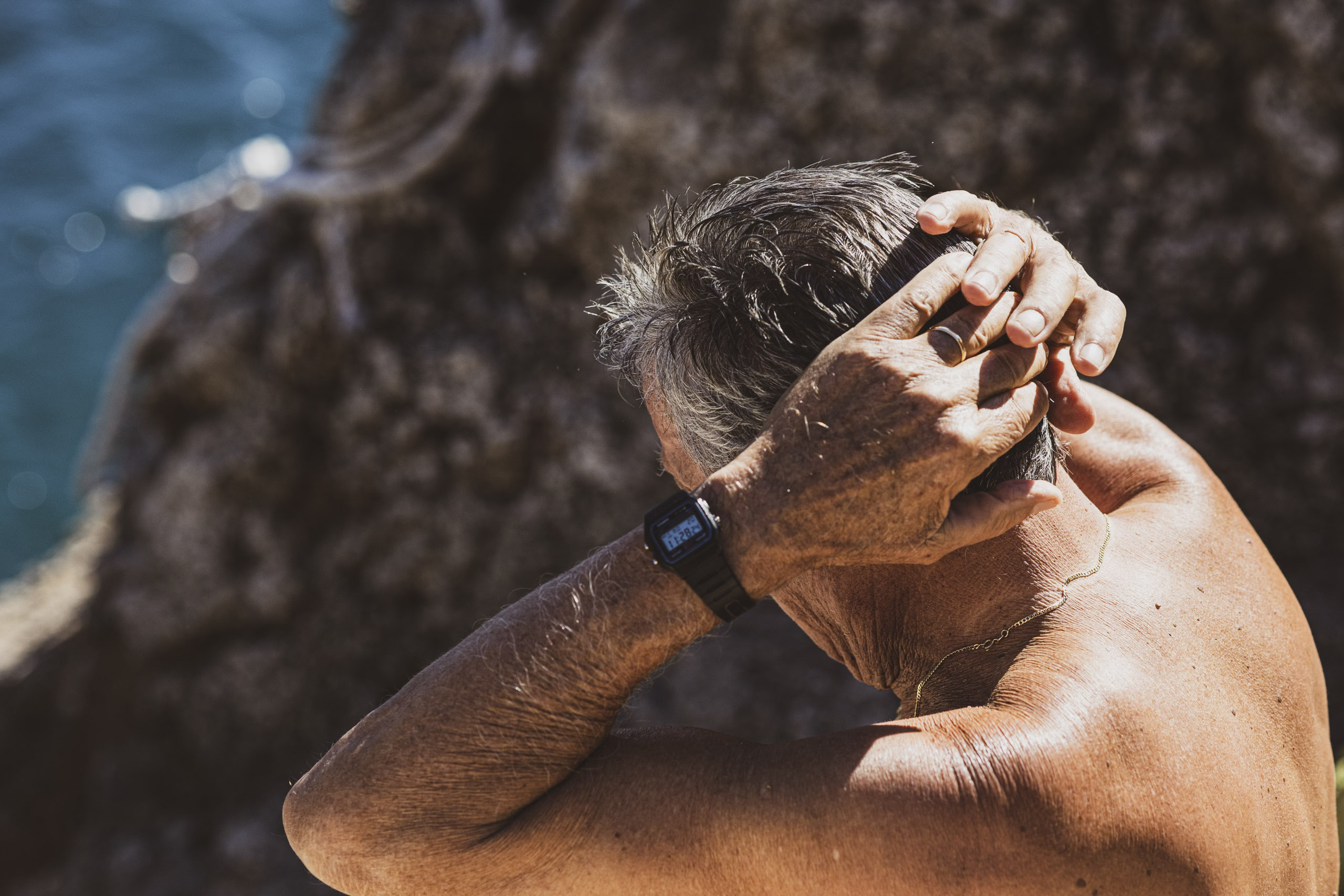 Ein älterer Mann mit graumeliertem Haar stellt am Meeresufer seine Uhr ein. Sein Körper ist sonnengebräunt und straff, im Hintergrund sind eine Felsklippe und das Meer unscharf. © Fotografie Tomas Rodriguez