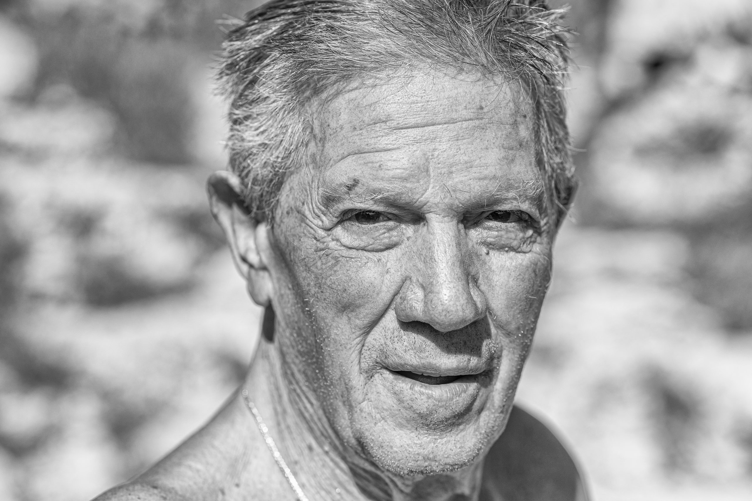 Nahaufnahme eines Schwarzweißfotos eines älteren Mannes mit nachdenklichem Gesichtsausdruck, wobei der Schwerpunkt auf seinen detaillierten Gesichtszügen und seiner strukturierten Haut vor einem unscharfen natürlichen Hintergrund liegt. © Fotografie Tomas Rodriguez