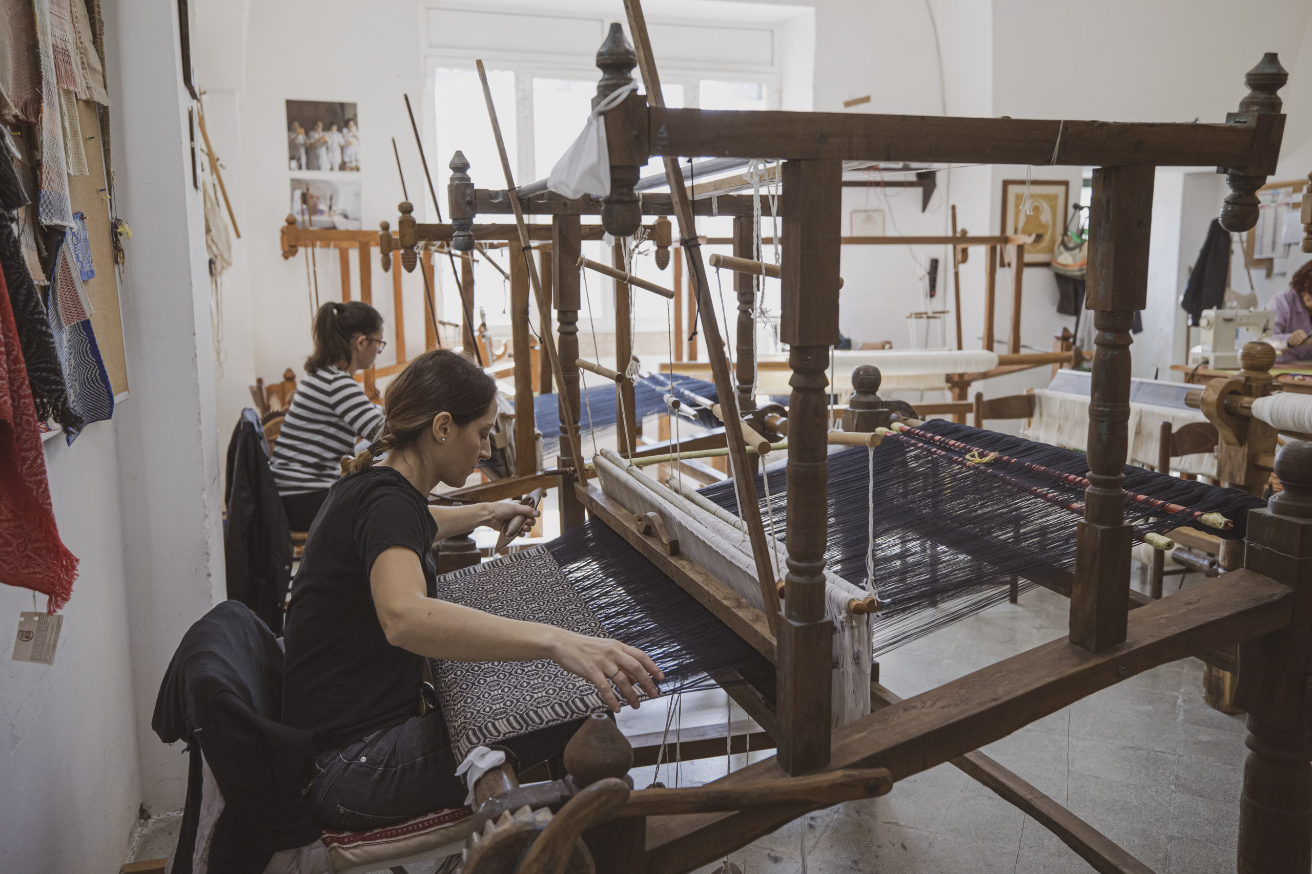 Eine Frau sitzt in einer Werkstatt an einem großen traditionellen Holzwebstuhl und webt Fäden. Im Hintergrund ist teilweise eine weitere Person zu sehen, die ebenfalls an einem Textilprojekt arbeitet. © Fotografie Tomas Rodriguez