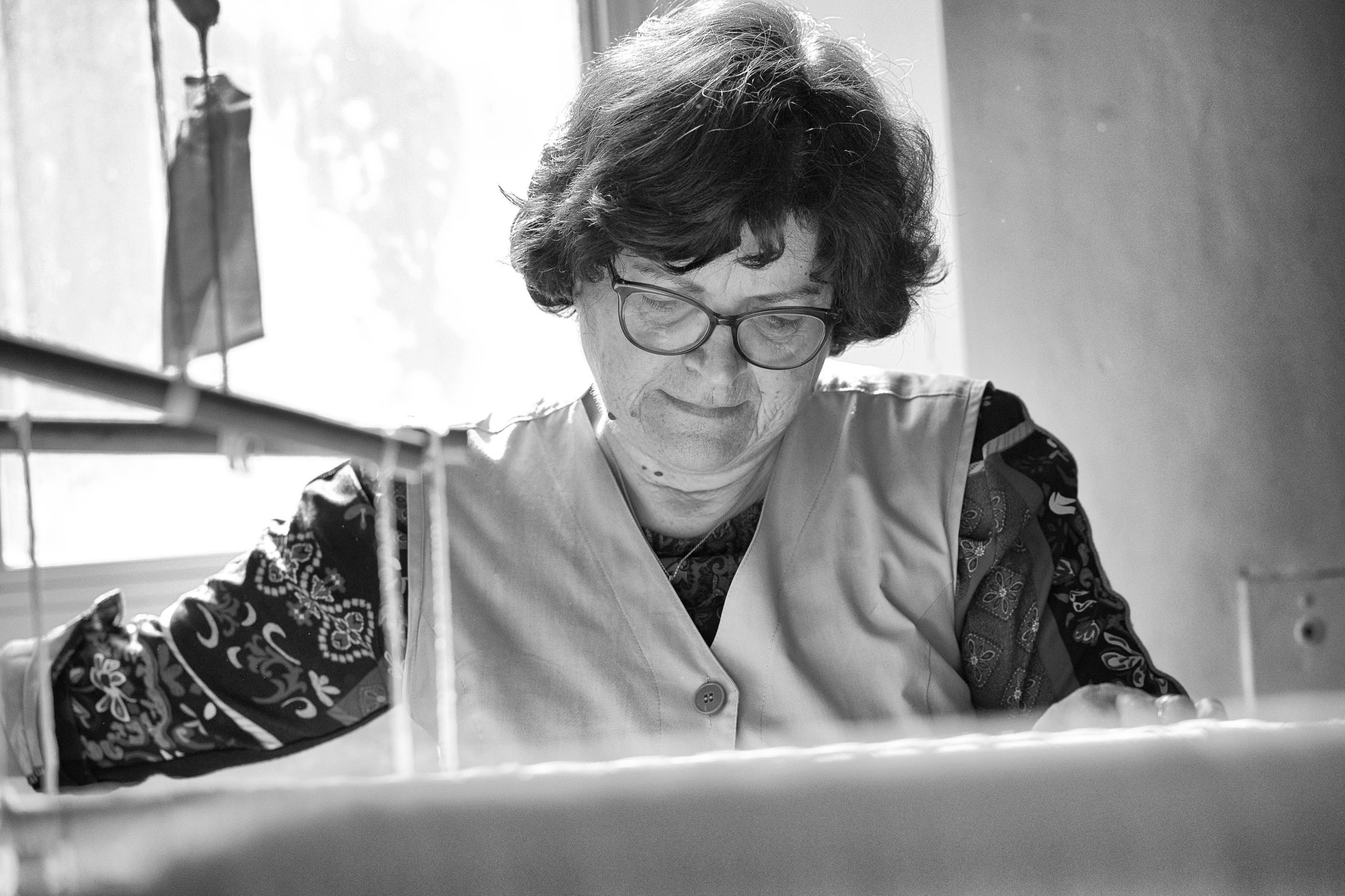 Eine ältere Frau mit Brille konzentriert sich intensiv auf ihre Arbeit an einem Schreibtisch, der vom Tageslicht eines nahegelegenen Fensters beleuchtet wird. Sie trägt eine gemusterte Bluse und eine Weste. © Fotografie Tomas Rodriguez
