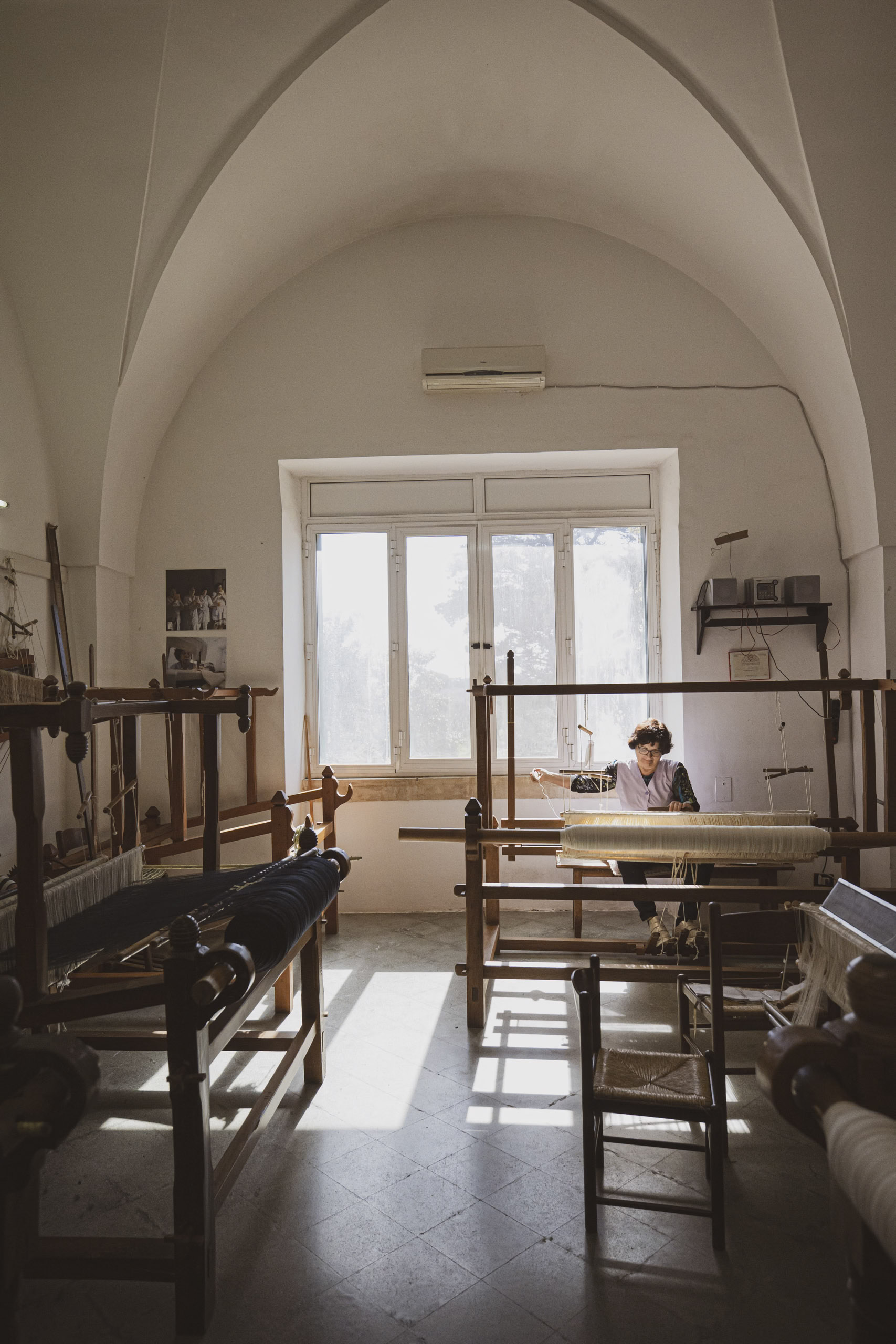 Frau webt an einem Webstuhl in einem sonnendurchfluteten, gewölbten Studio mit mehreren Webstühlen und einer friedlichen, künstlerischen Atmosphäre. © Fotografie Tomas Rodriguez