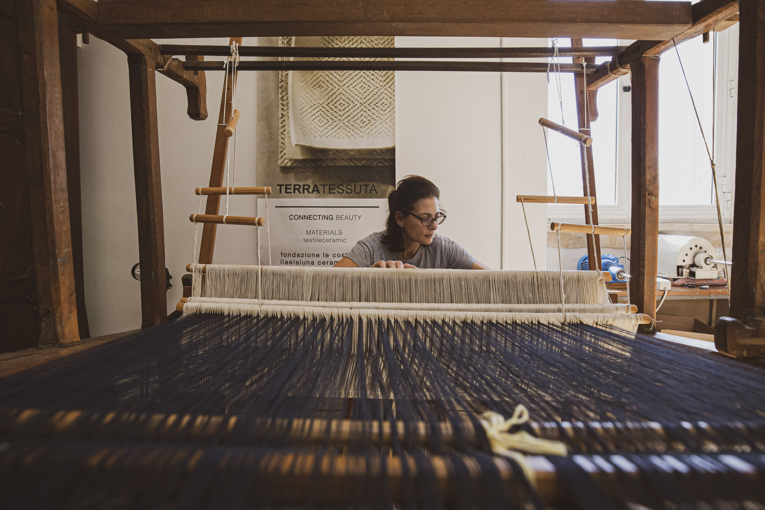 Eine Frau webt in einer warm beleuchteten Werkstatt an einem großen Holzwebstuhl und konzentriert sich dabei auf die Reihen aus weißen und blauen Fäden. Darüber hängt ein Stoff mit komplizierten Mustern. © Fotografie Tomas Rodriguez