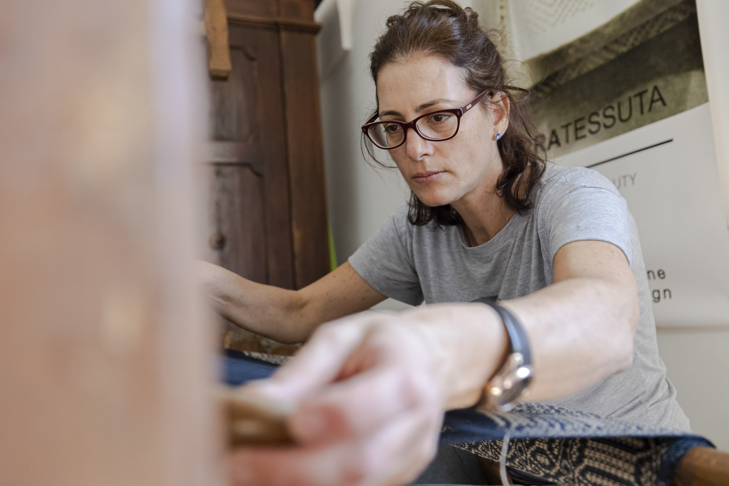 Eine konzentrierte Frau mit Brille und grauem T-Shirt schleift mit einem Schleifblock eine Holzoberfläche und arbeitet konzentriert an einem Projekt im Innenbereich. © Fotografie Tomas Rodriguez