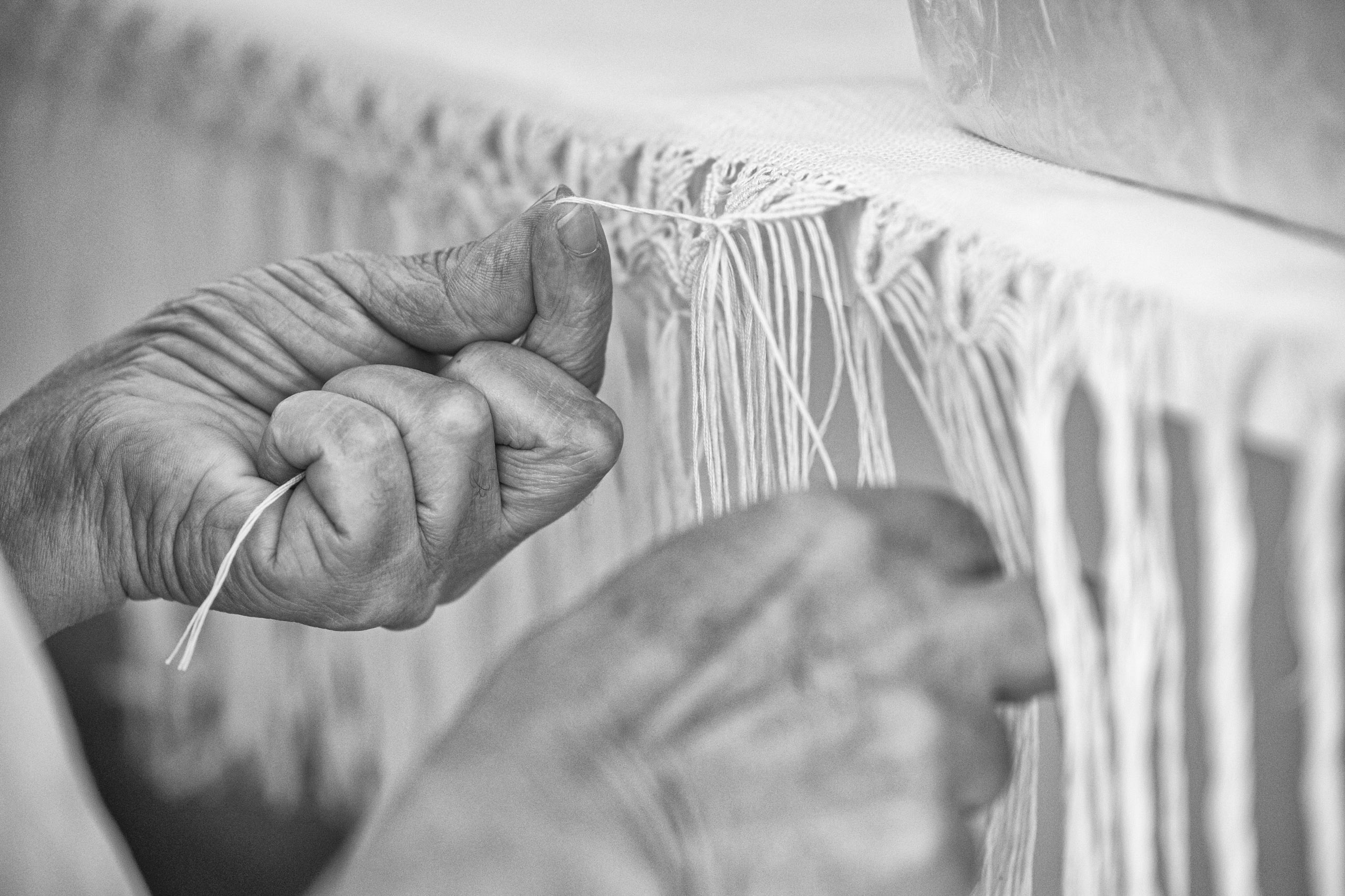Nahaufnahme älterer Hände, die geschickt Fransen in einen Stoff binden, wobei der Schwerpunkt auf der präzisen Bewegung und der zarten Berührung in einer monochromen Umgebung liegt. © Fotografie Tomas Rodriguez