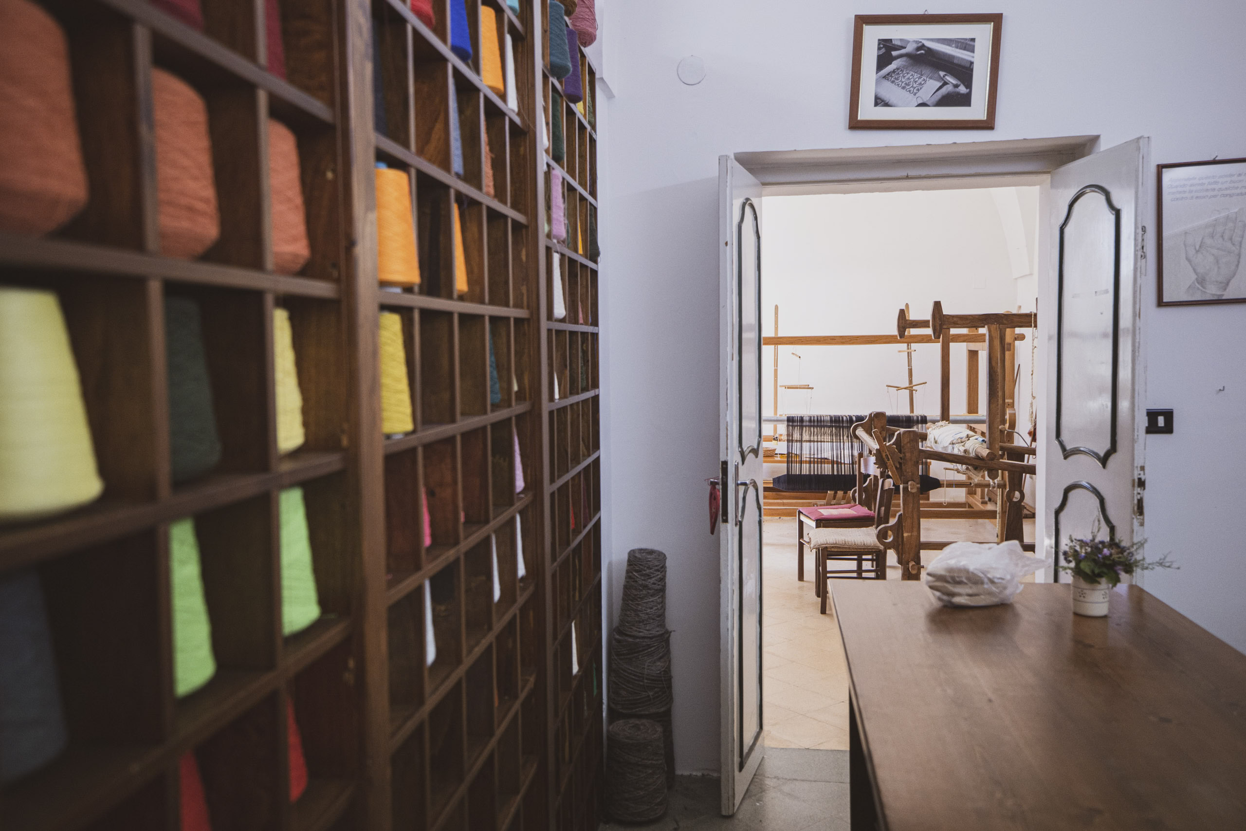 Eine Textilwerkstatt mit einem großen Regal voller bunter Garnspulen auf der linken Seite, das zu einem Raum mit einem hölzernen Webstuhl und Bilderrahmen an den Wänden führt. © Fotografie Tomas Rodriguez