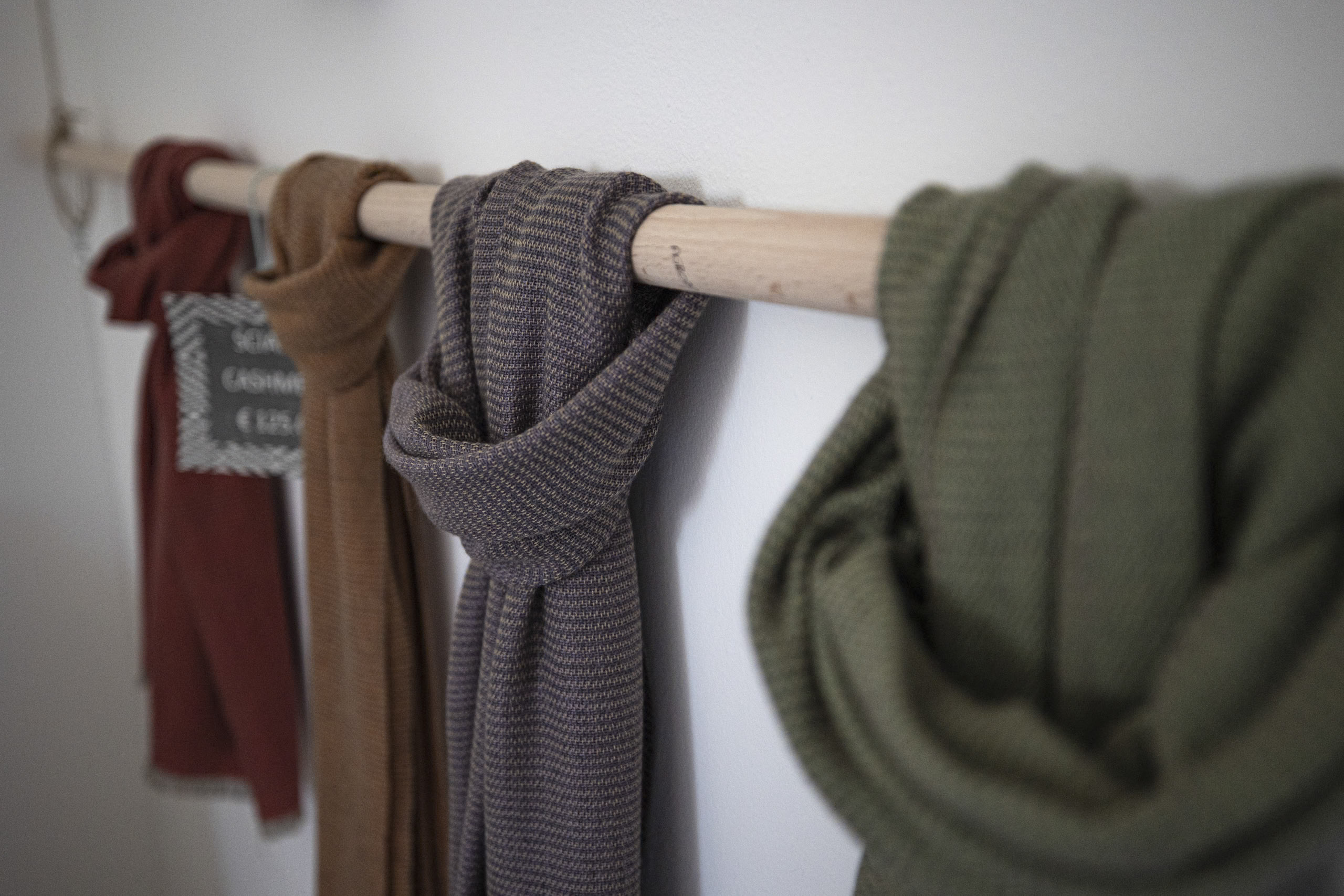 Eine an einer weißen Wand montierte Garderobe aus Holz, an der mehrere Schals und eine Strickjacke lose hängen. Der Fokus liegt auf einem grau karierten Schal, wobei der Hintergrund sanft unscharf ist. © Fotografie Tomas Rodriguez