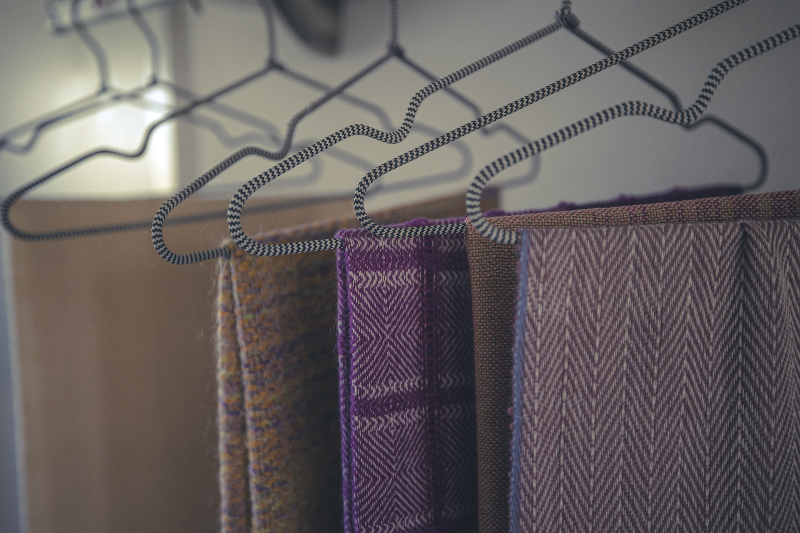 Nahaufnahme mehrerer Tweedjacken, die auf stilvollen Metallbügeln vor einem weichgezeichneten Hintergrund hängen, wodurch die Texturen und Muster der Stoffe hervorgehoben werden. © Fotografie Tomas Rodriguez