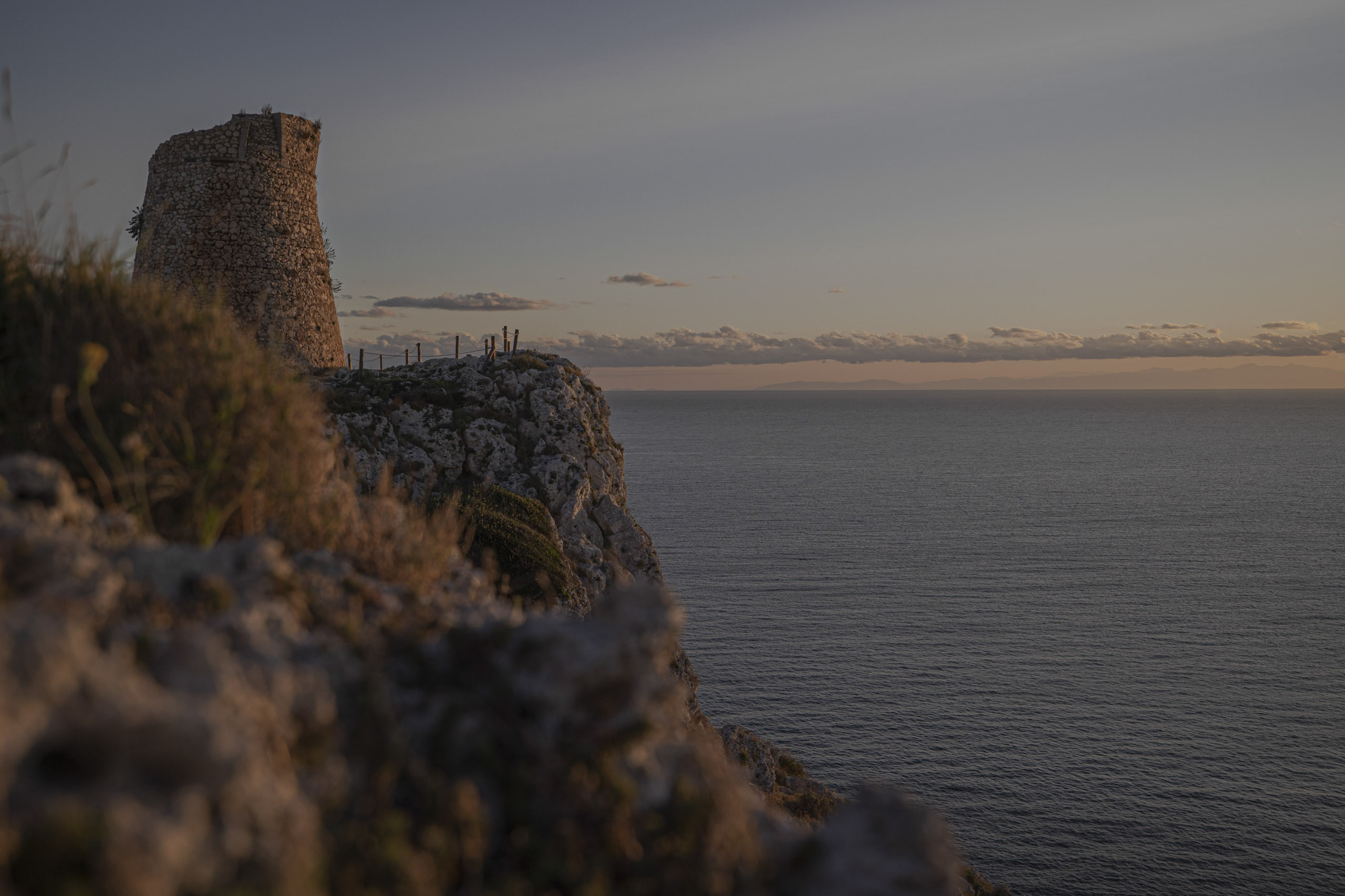 Ein alter Steinturm thront auf einer schroffen Klippe und überblickt bei Sonnenuntergang das Meer. Sanftes, goldenes Licht wirft einen warmen Farbton auf die Szene. © Fotografie Tomas Rodriguez