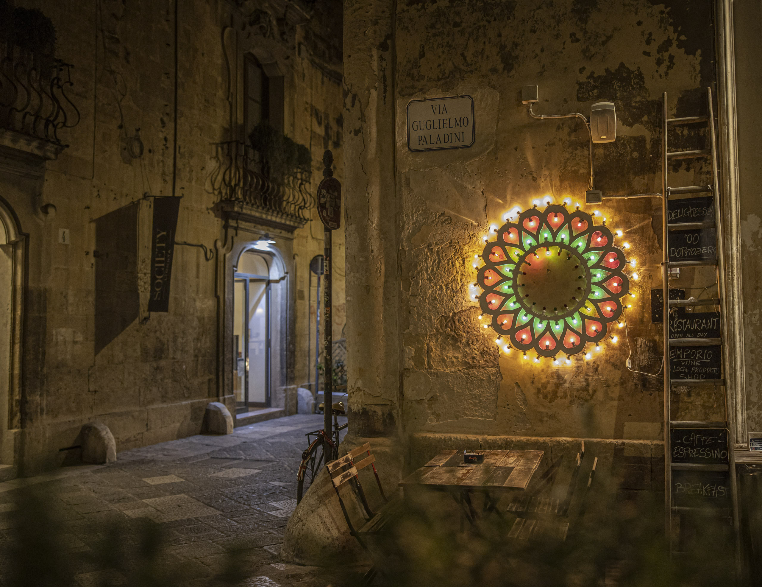 Ein leuchtendes, rundes Neonschild erhellt nachts eine gemütliche Straßenecke in einer alten europäischen Stadt mit Kopfsteinpflasterstraßen und malerischen Gebäuden, die mit Balkonen und Straßenschildern geschmückt sind. © Fotografie Tomas Rodriguez