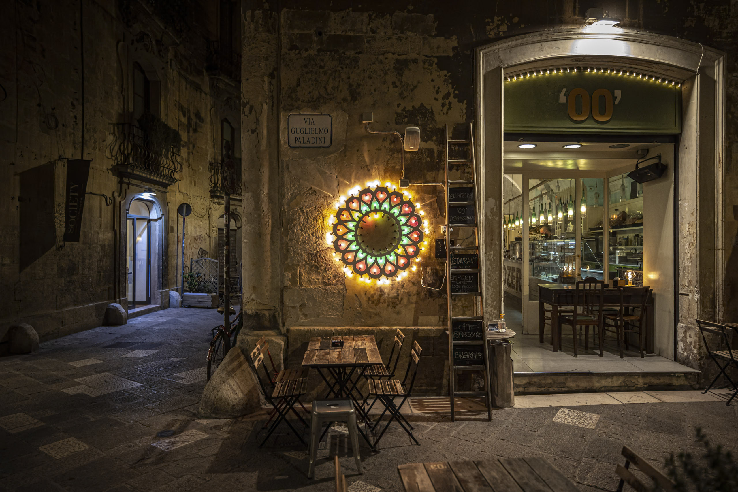 Ein charmantes Café bei Nacht in einer alten Kopfsteinpflastergasse, dekoriert mit einer lebendigen runden Lichtskulptur, einladenden Holztischen im Freien und warmer Innenbeleuchtung. © Fotografie Tomas Rodriguez