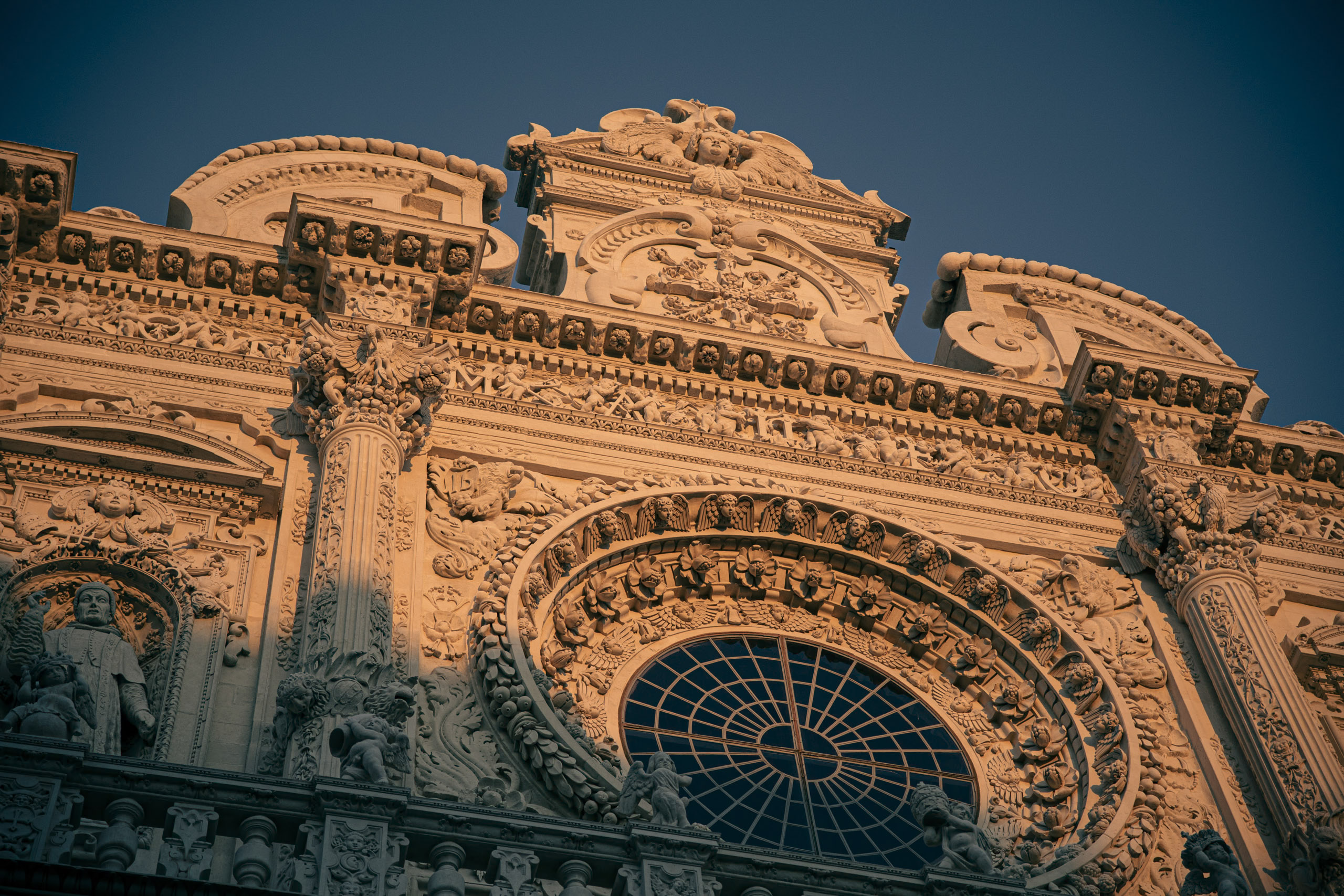 Aufwändiges architektonisches Detail eines historischen Gebäudes mit aufwendigen Schnitzereien und einem großen, detaillierten runden Fenster, das in warmes Licht getaucht ist. © Fotografie Tomas Rodriguez