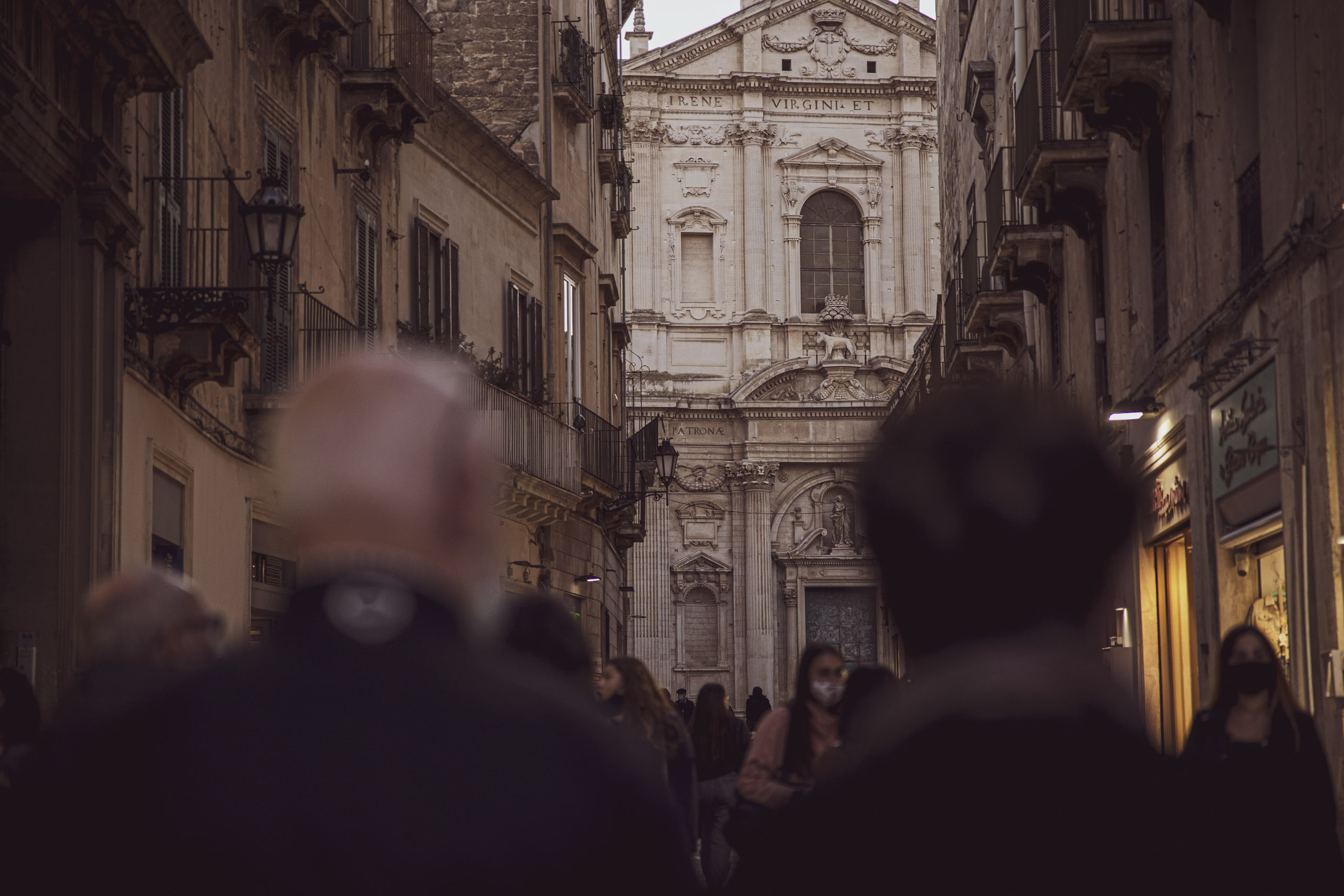 Blick auf eine belebte Straße, die zu einer historischen Kirche mit einer detailreichen Fassade führt. Menschen mit Masken gehen entlang, was auf Vorsichtsmaßnahmen gegen die Pandemie hinweist. Die Szene fängt eine Mischung aus Architektur und Alltag ein. © Fotografie Tomas Rodriguez