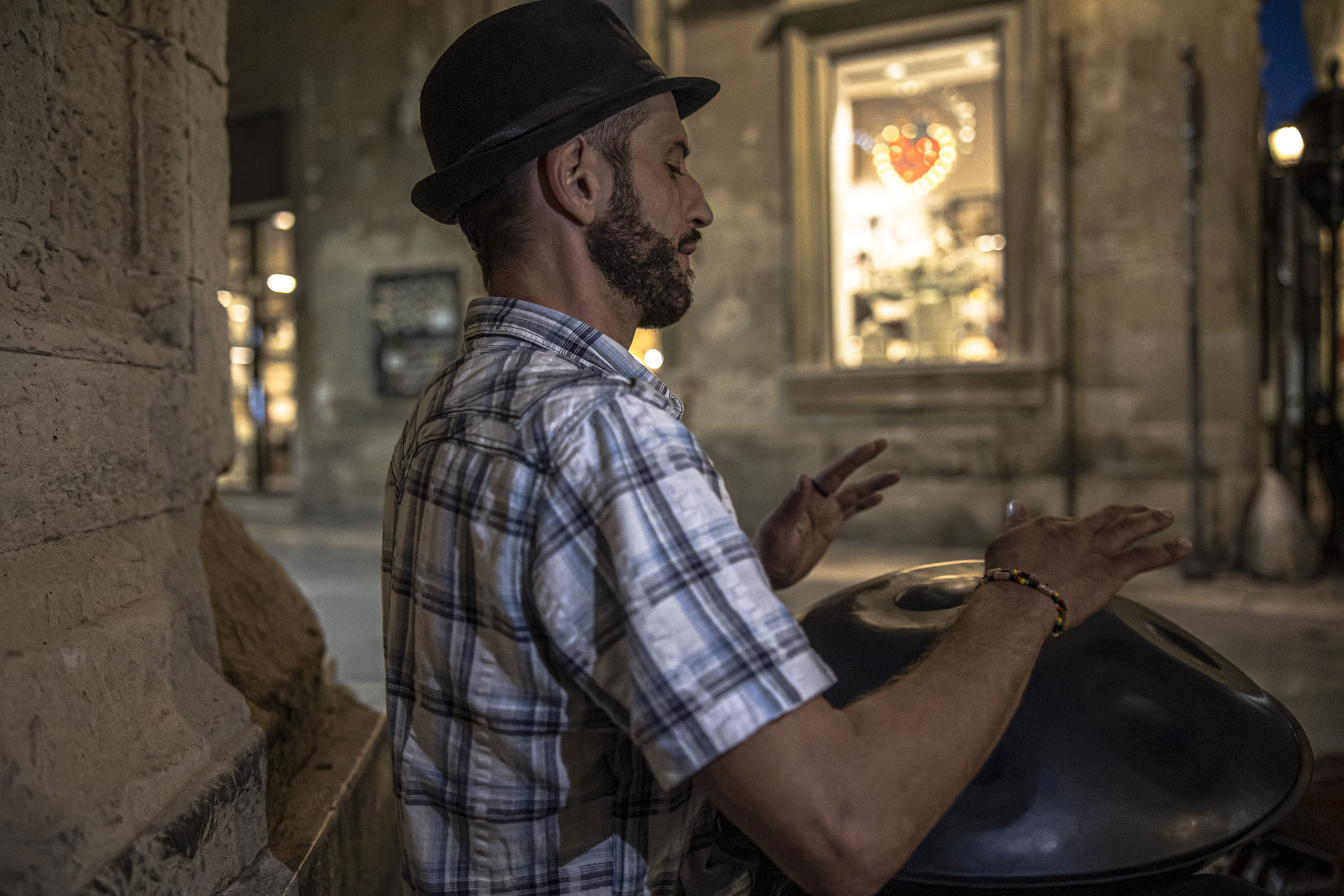 Ein bärtiger Mann mit Fedora spielt nachts auf einer historischen Straße eine Hang-Trommel. Sein konzentrierter Gesichtsausdruck wird durch das Umgebungslicht eines Schaufensters hinter ihm beleuchtet. © Fotografie Tomas Rodriguez