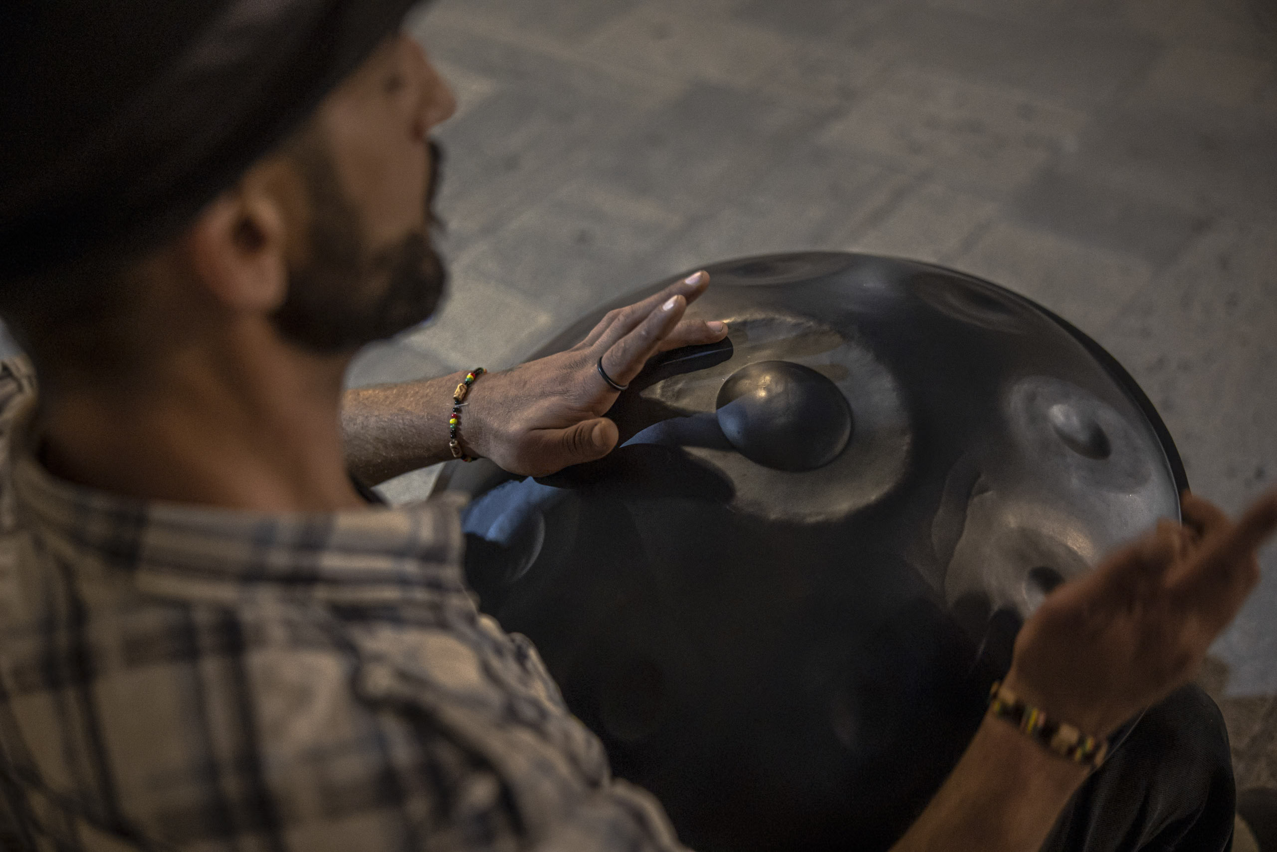 Ein Mann in kariertem Hemd und Hut spielt ein Handpan-Instrument aus Stahl und klopft sanft mit seinen Fingern auf die geschwungenen Vertiefungen. Der Fokus liegt auf seinen Händen auf dem Instrument in einer schwach beleuchteten Umgebung. © Fotografie Tomas Rodriguez