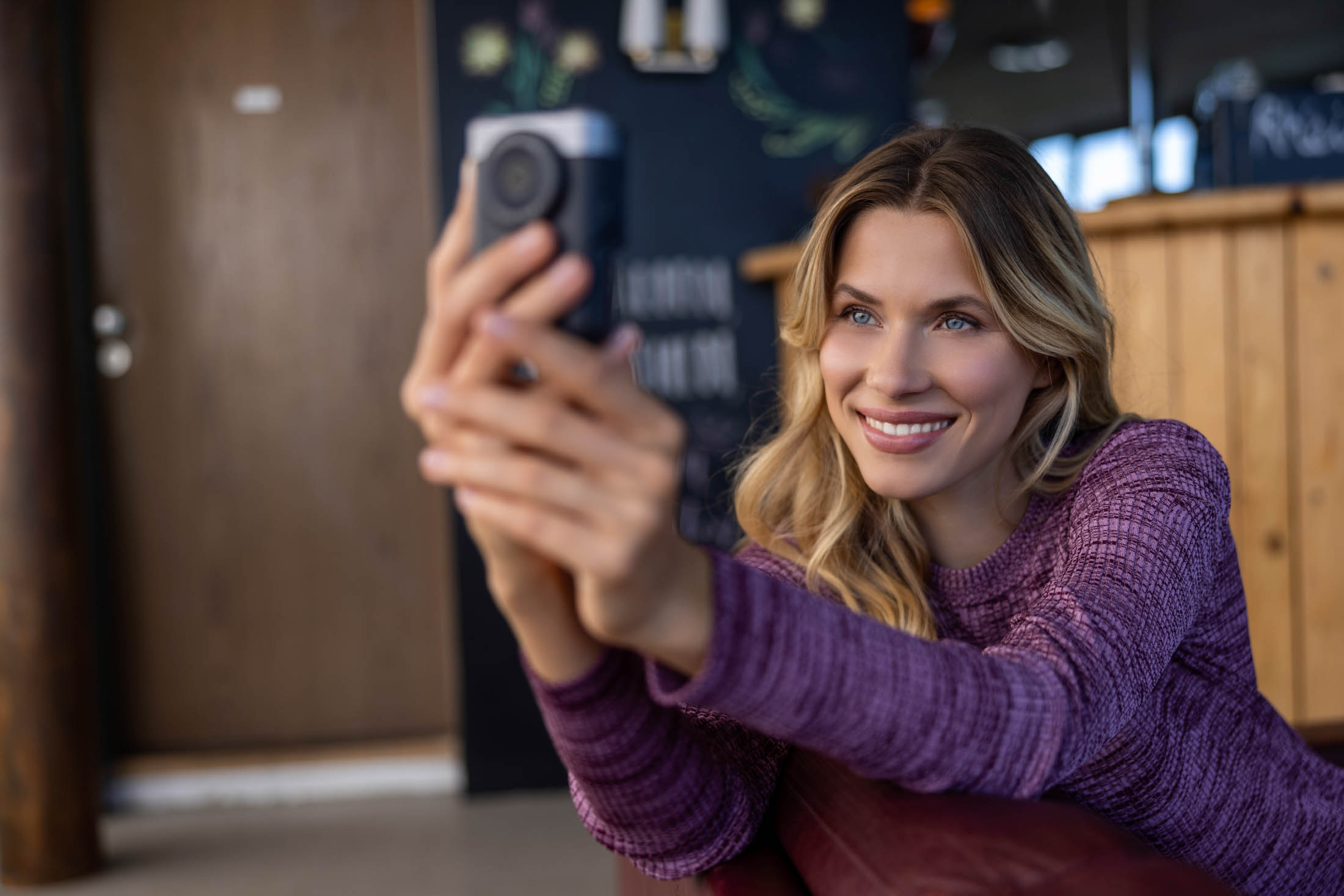 Eine lächelnde Frau im lila Pullover macht in einem gemütlichen Café-Ambiente ein Selfie mit ihrem Smartphone. © Fotografie Tomas Rodriguez