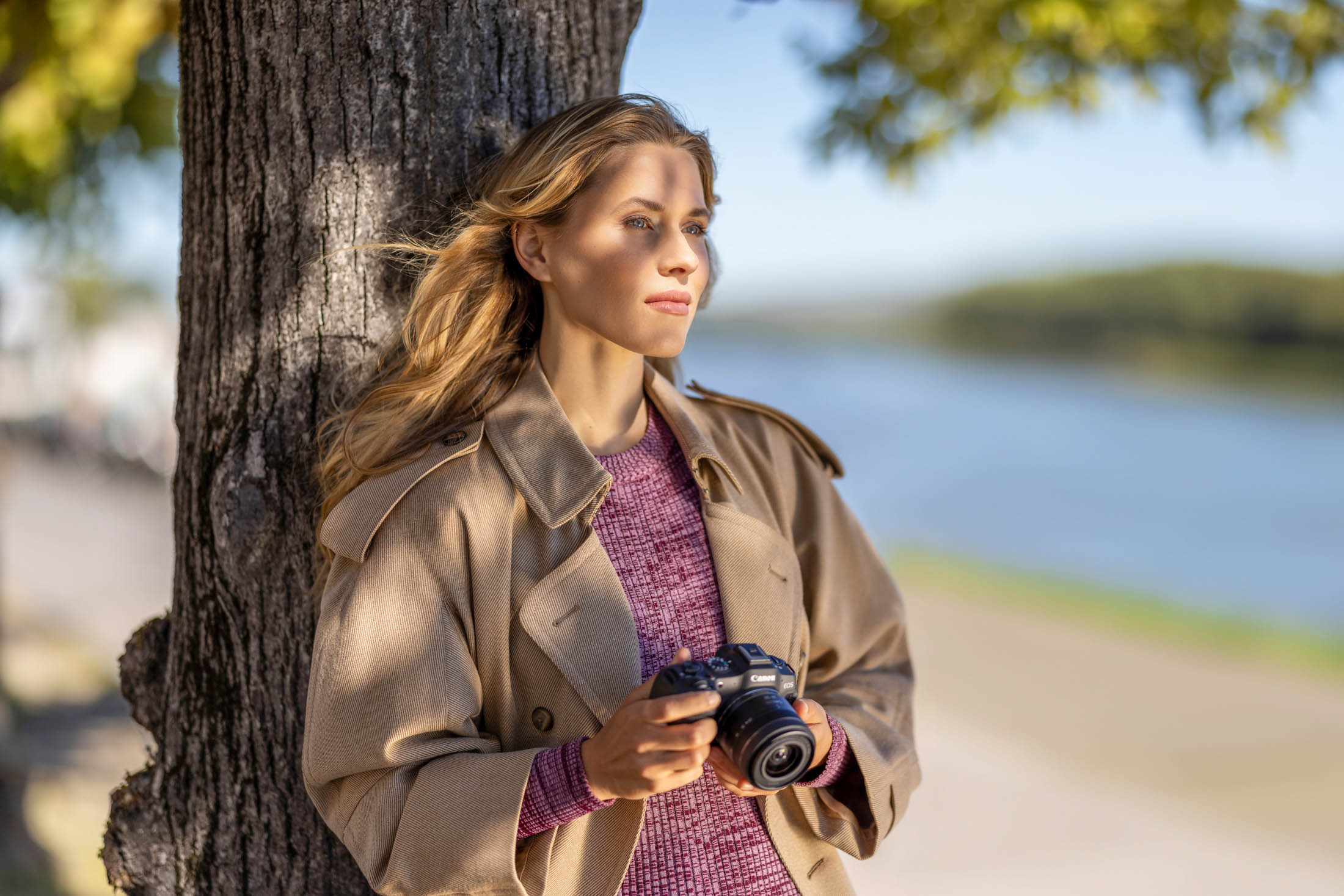 Eine nachdenkliche Frau lehnt mit einer Kamera in der Hand an einem Baum am Fluss. Sie trägt einen beigen Trenchcoat über einem lila Pullover, ihr Haar weht sanft im Wind und sie ist in tiefes Nachdenken versunken. For Canon Deutschland © Fotografie Tomas Rodriguez