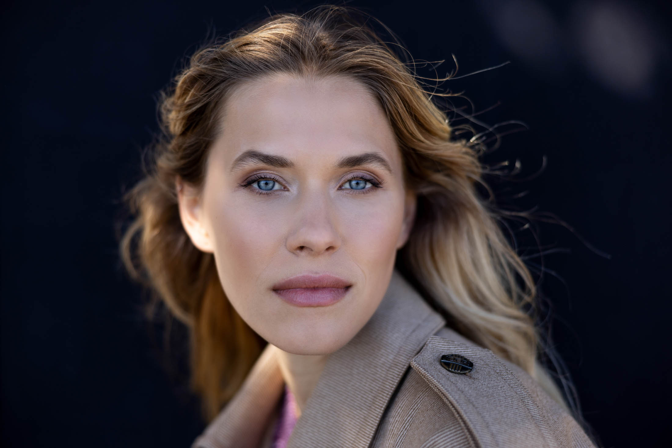 Ein Nahaufnahmeporträt einer Frau mit langen blonden Haaren, klaren blauen Augen und leichtem Make-up, die einen beigen Mantel trägt und vor einem unscharfen schwarzen Hintergrund aufmerksam in die Kamera blickt. © Fotografie Tomas Rodriguez