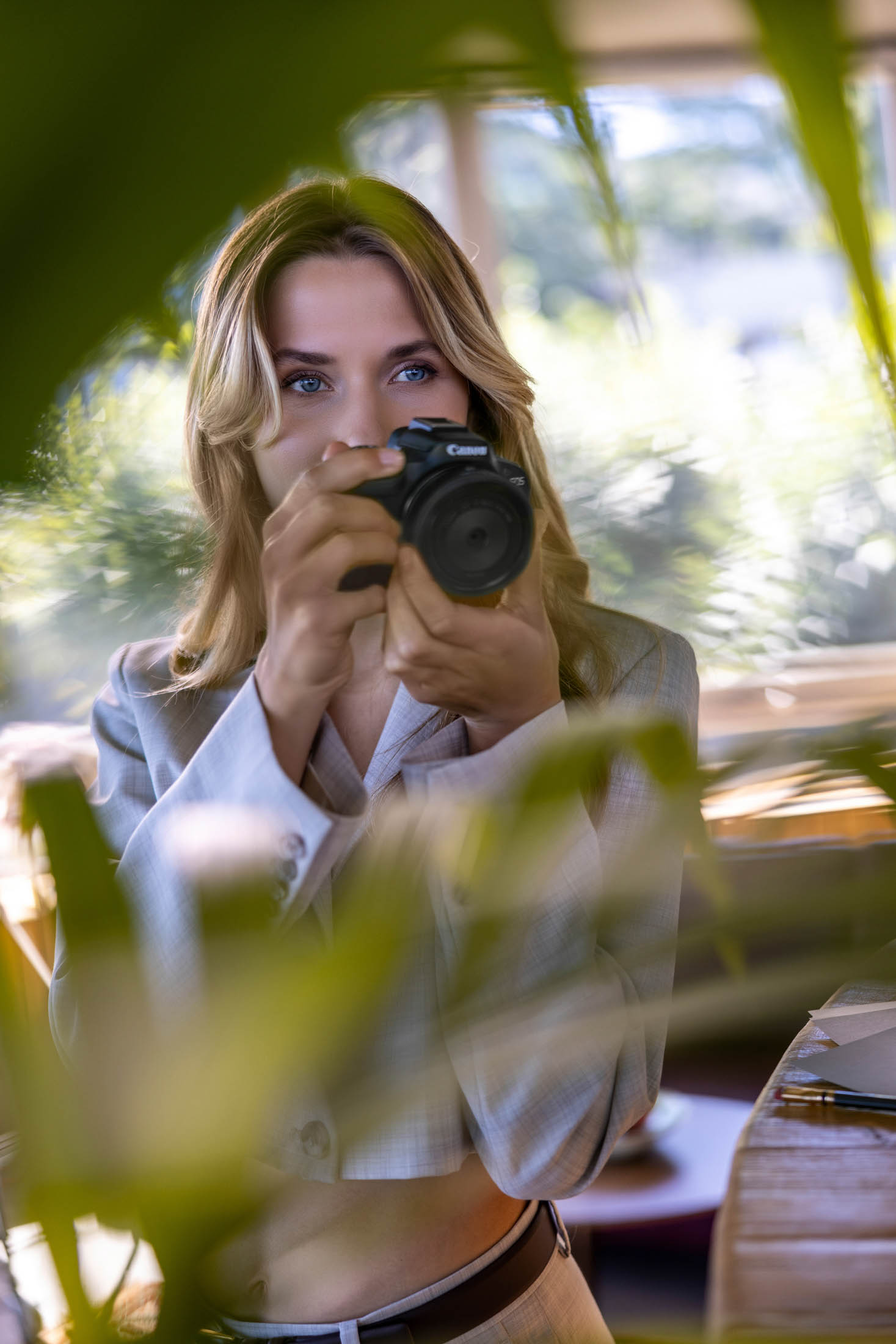 Eine Frau mit blondem Haar hält eine Canon-Kamera in der Hand und ist bereit, ein Foto zu schießen. Sie lugt durch das grüne Laub in einem gut beleuchteten Raum. Sie trägt einen Business-Anzug und hat konzentrierte, aufmerksame blaue Augen. © Fotografie Tomas Rodriguez