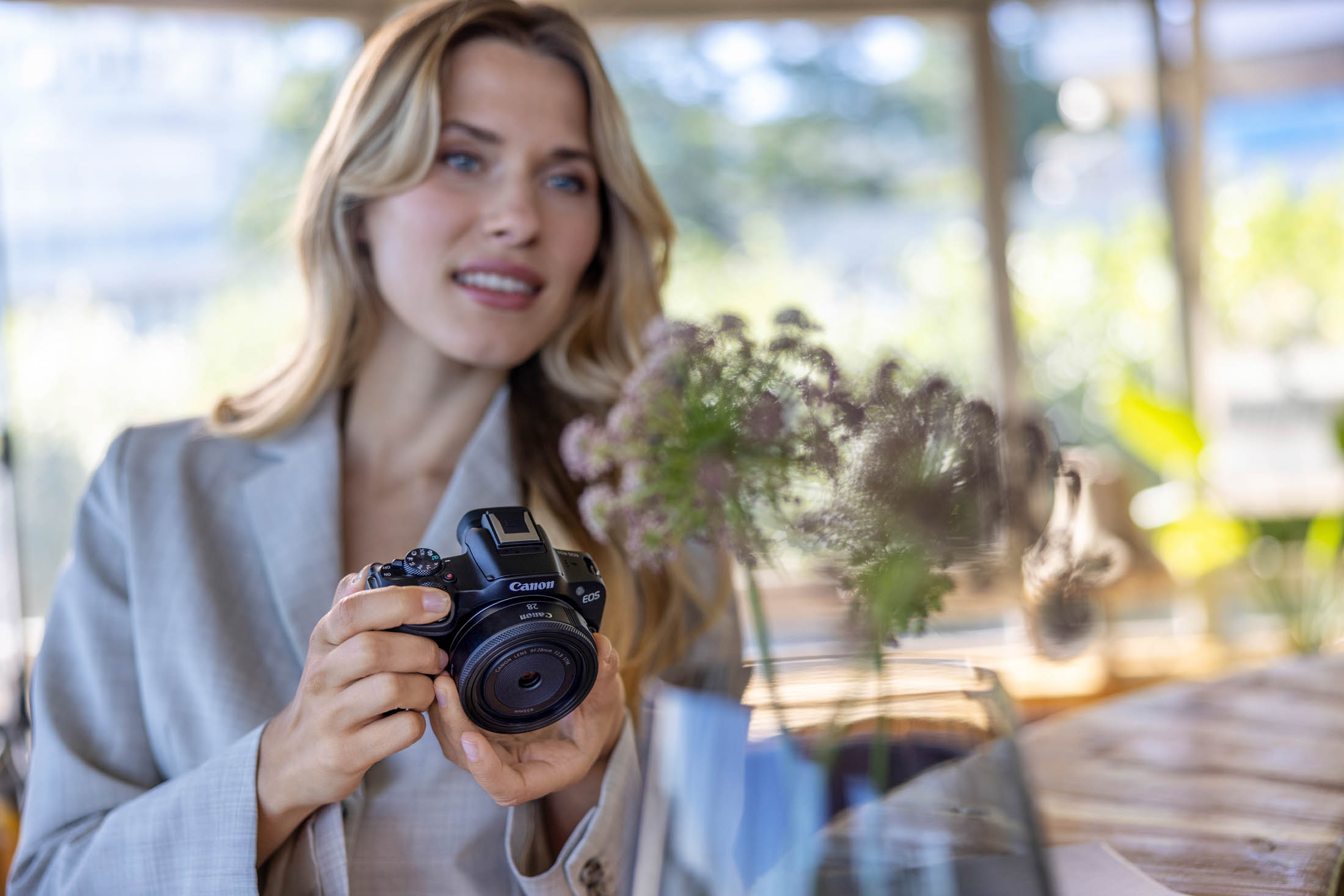Eine Frau hält eine Canon-Kamera und fokussiert intensiv auf ihr Motiv. Im unscharfen Hintergrund ist ein helles, luftiges Café mit Pflanzen zu sehen. Sie scheint sich mit Fotografieren zu beschäftigen. © Fotografie Tomas Rodriguez