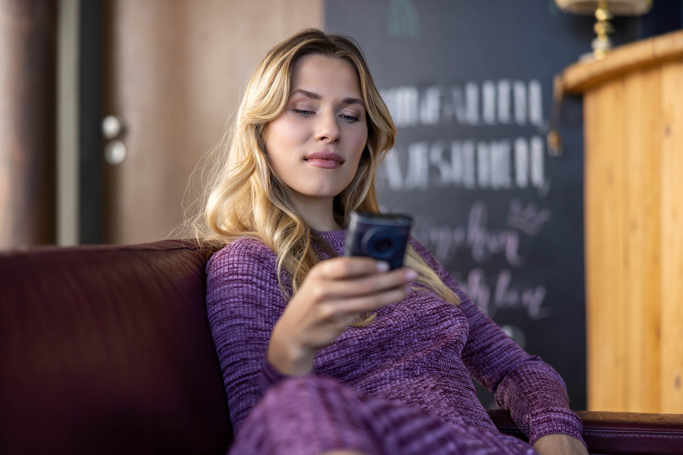 Eine Frau in einem lila Pullover sitzt gemütlich auf einem Sofa und schaut auf ihr Smartphone, vor dem Hintergrund eines stilvollen Zimmers mit weichem Fokus. © Fotografie Tomas Rodriguez