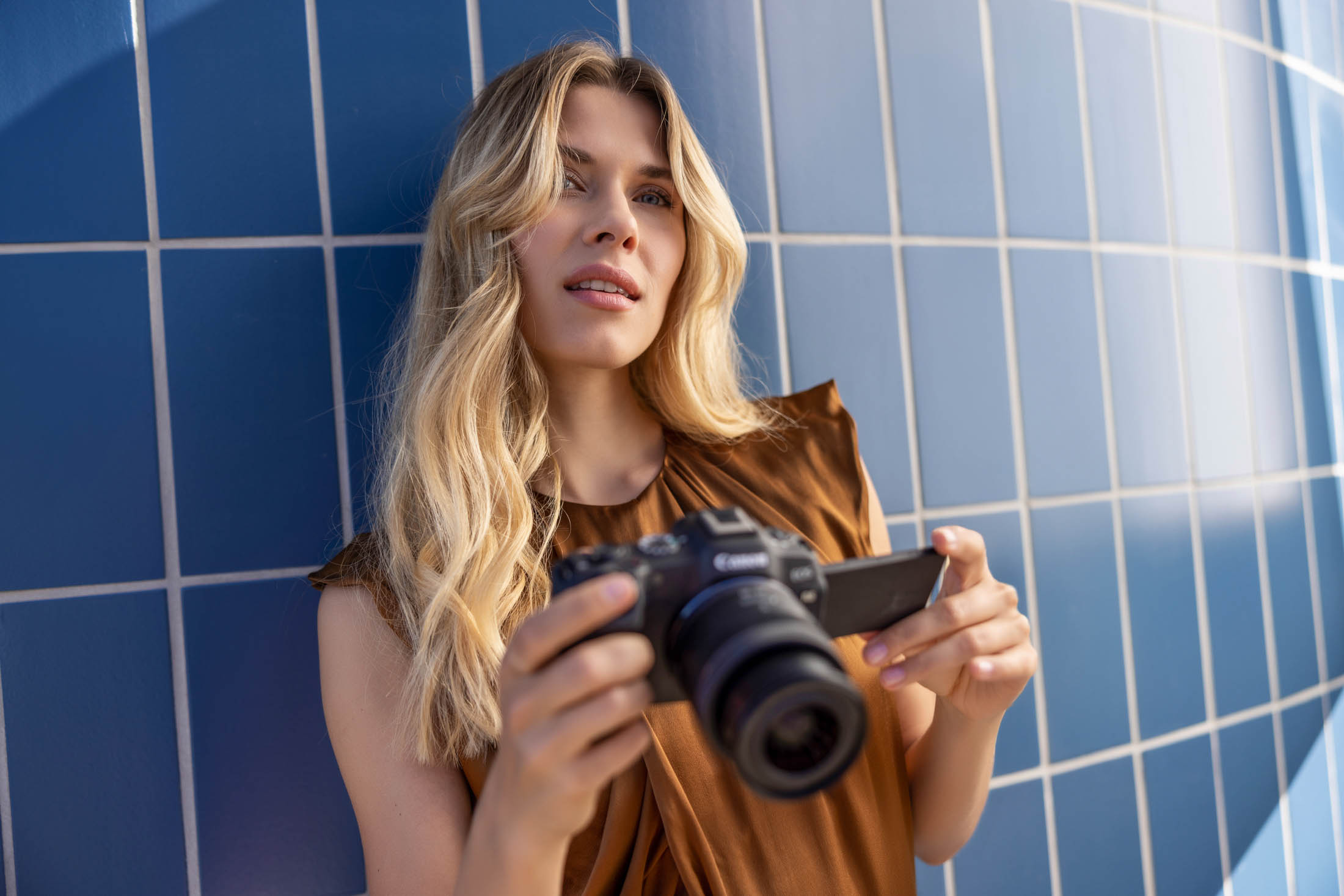 Canon, Rheinpavillion Bonn. Eine Frau mit blondem Haar betrachtet Bilder auf einer Digitalkamera und steht vor einer blau gefliesten Wand. Sie trägt ein ärmelloses Top und sieht nachdenklich aus. © Fotografie Tomas Rodriguez