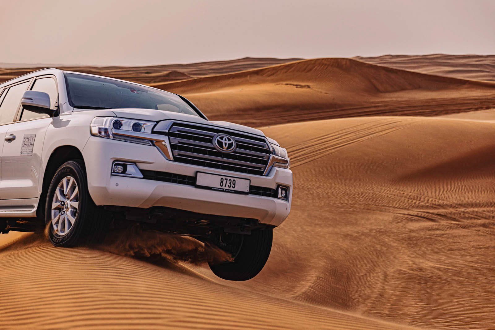 Ein weißer Toyota Land Cruiser fährt über eine Sanddüne in einer Wüstenlandschaft, eine Staubwolke hinterlässt eine Spur, das Nummernschild ist sichtbar. © Fotografie Tomas Rodriguez