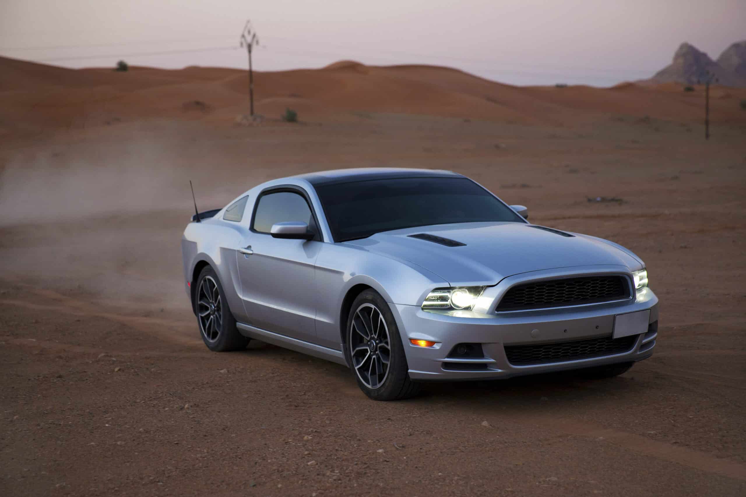 Ein silberner Ford Mustang parkt in der Dämmerung auf einer staubigen Wüstenstraße, mit sanften Orangetönen am Himmel und Sanddünen im Hintergrund. © Fotografie Tomas Rodriguez