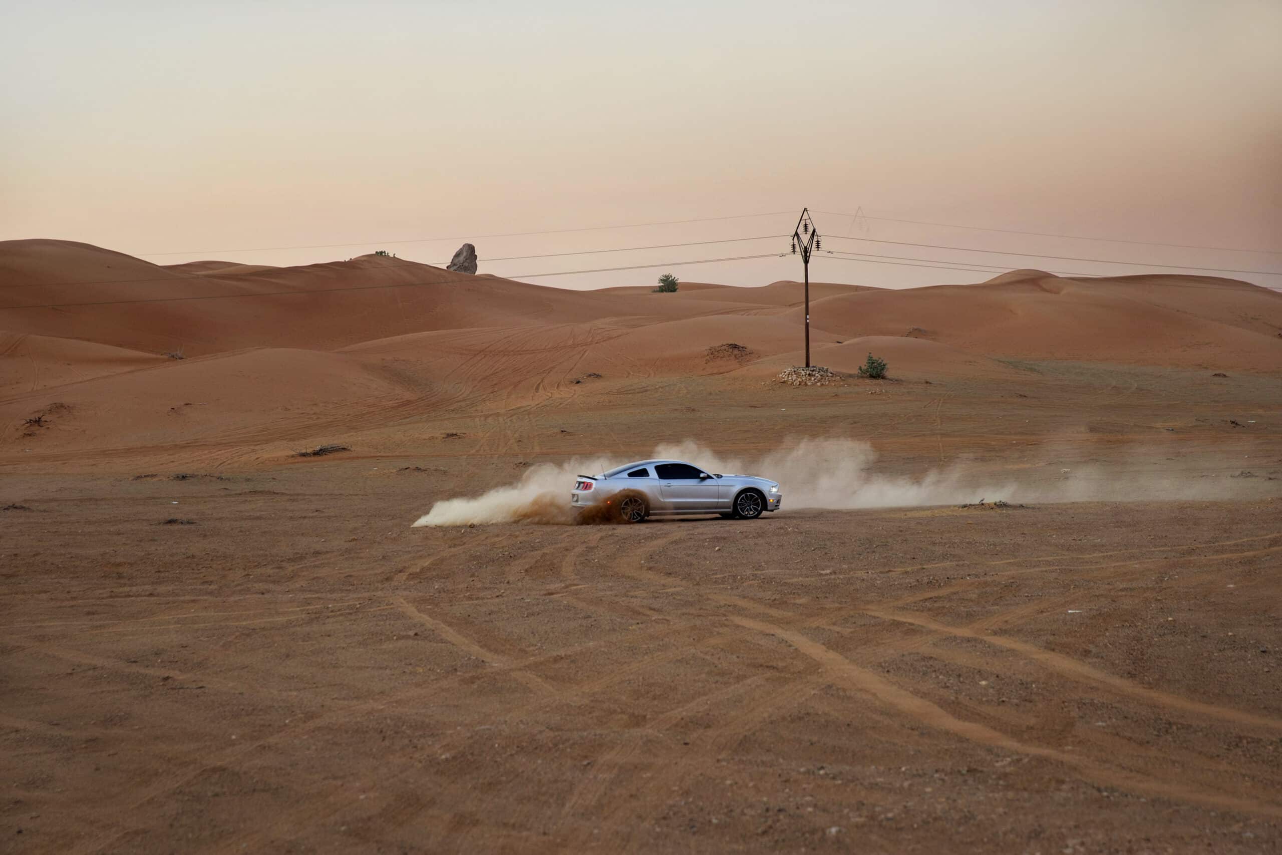 Ein silberner Sportwagen rast durch eine Wüstenlandschaft und wirbelt unter einem sanften Sonnenuntergangshimmel Staub auf. Im Hintergrund sind spärliche Vegetation und sanfte Sanddünen zu sehen. © Fotografie Tomas Rodriguez