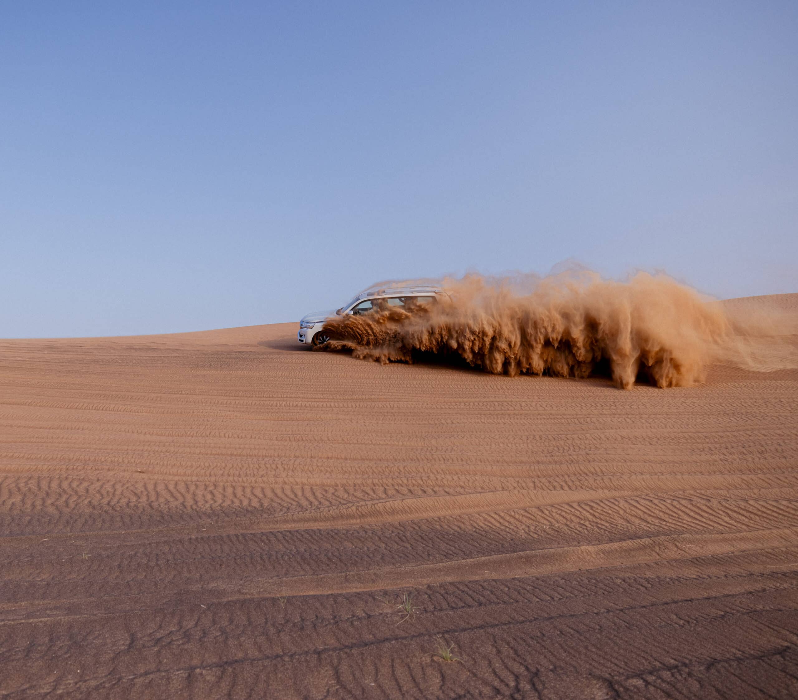 Ein silberner SUV wirbelt eine große Sandwolke auf, während er unter einem klaren blauen Himmel schnell durch eine weite Wüstenlandschaft fährt. © Fotografie Tomas Rodriguez