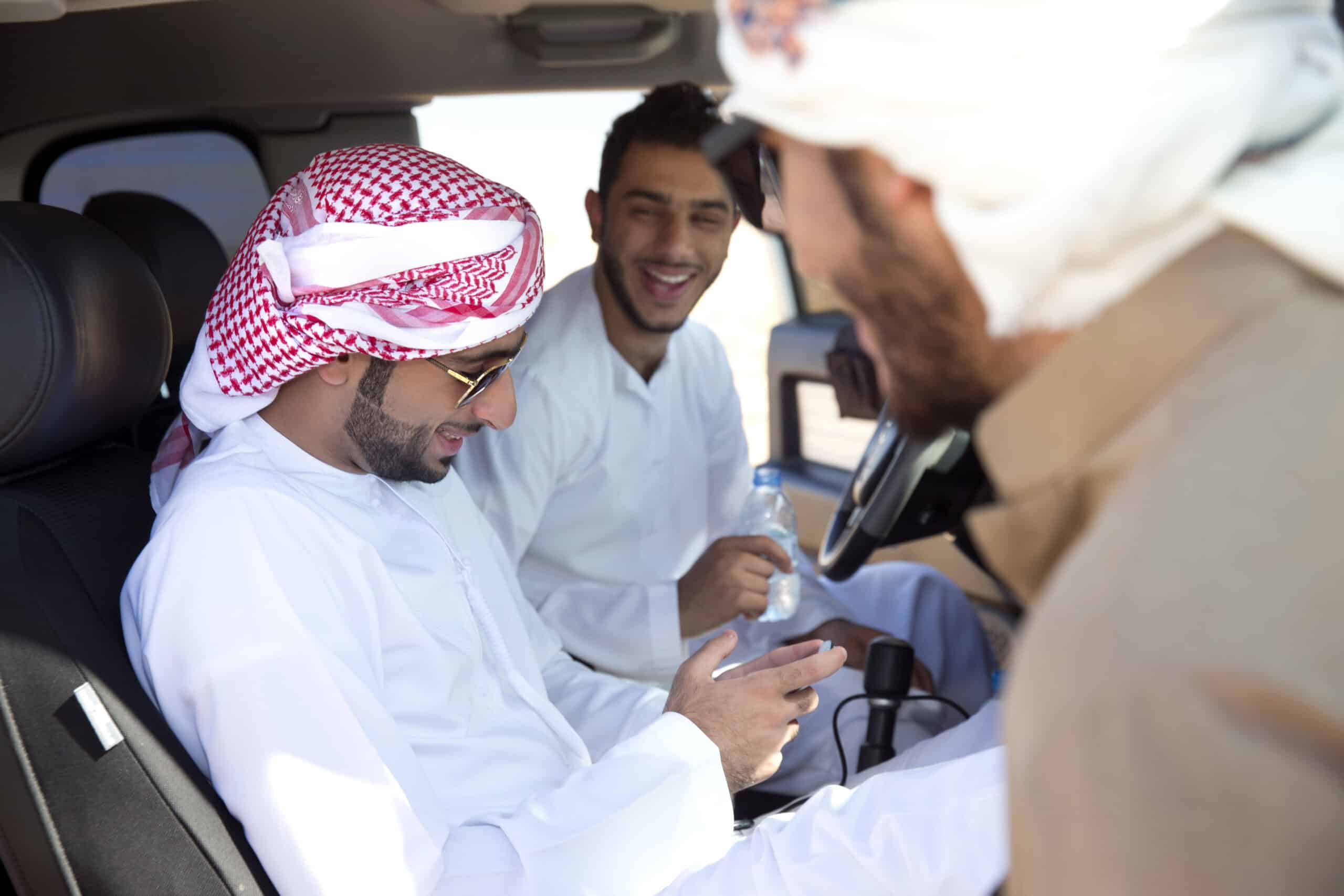 Drei Männer in traditioneller arabischer Kleidung lächeln und unterhalten sich in einem Auto; zwei schauen auf das Smartphone, das einer von ihnen in der Hand hält. © Fotografie Tomas Rodriguez