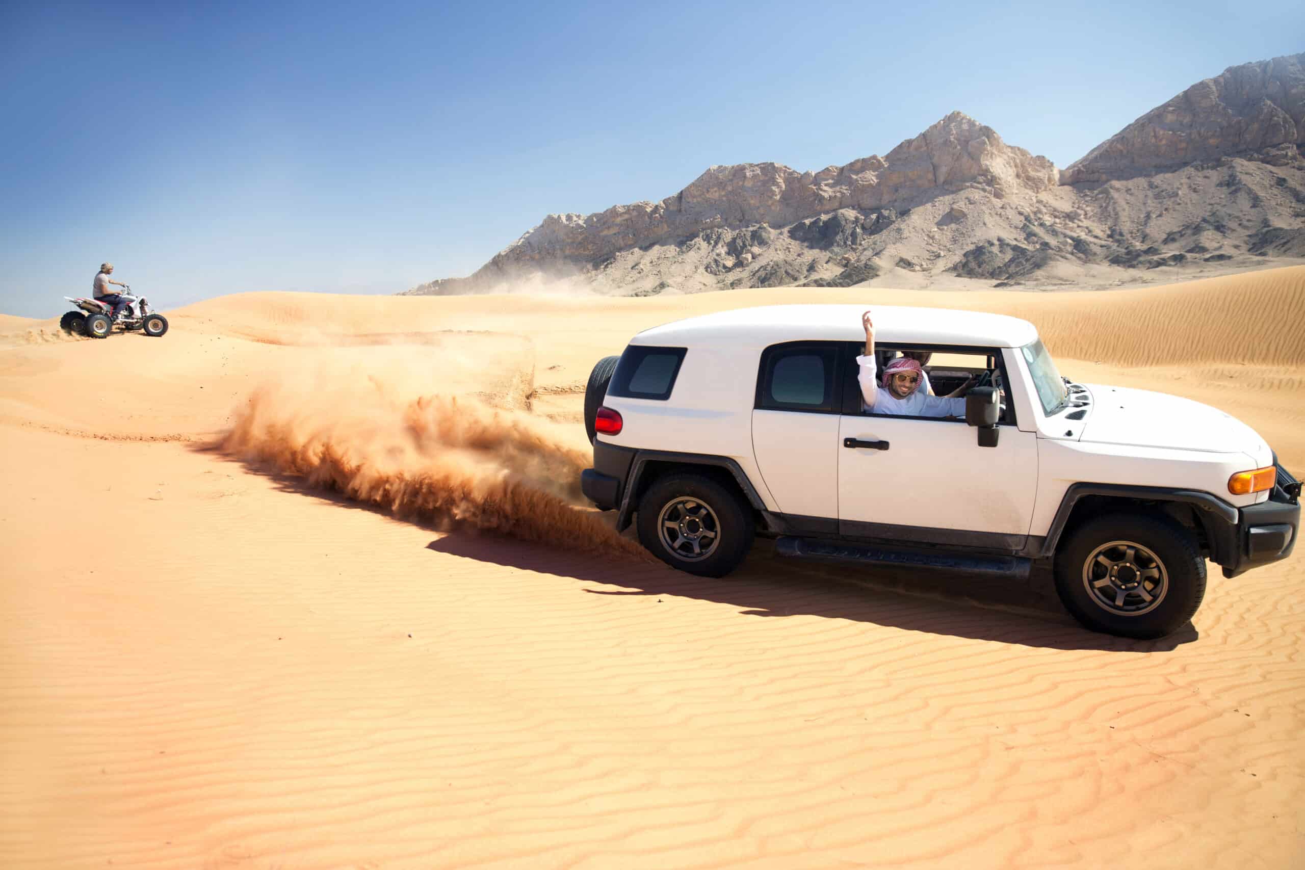 Ein Mann mit Sonnenbrille winkt aus einem weißen Geländewagen, der durch eine Sandwüste rast und Staub aufwirbelt. Im Hintergrund ist in der Nähe der Rocky Mountains eine Person auf einem Geländefahrzeug zu sehen. © Fotografie Tomas Rodriguez