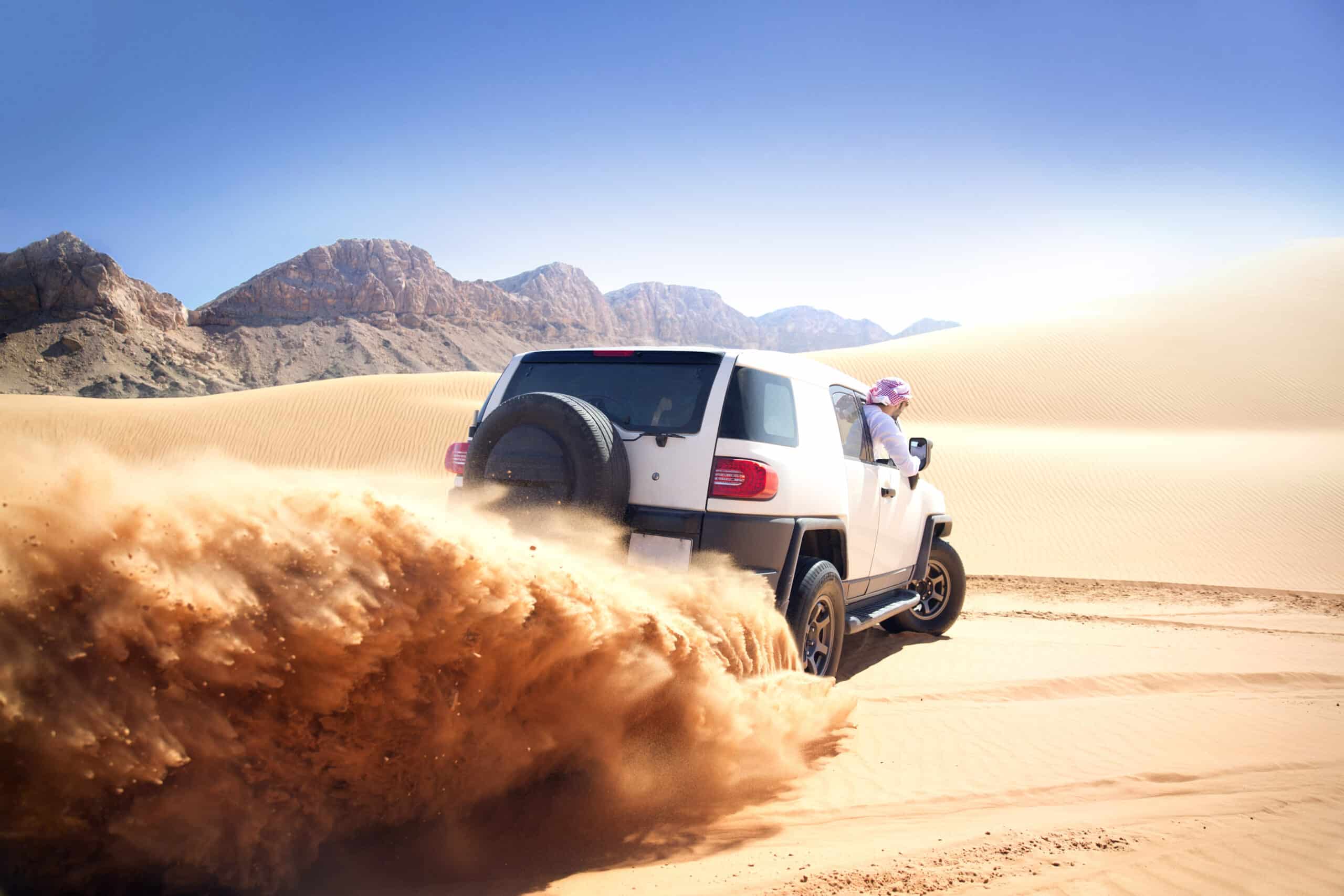 Ein weißer Geländewagen rast durch eine sandige Wüstenlandschaft und wirbelt dabei Sand auf. Im Hintergrund sind Berge zu sehen, der Himmel ist klar und blau. Aus dem Beifahrerfenster blickt eine Person mit Kopftuch. © Fotografie Tomas Rodriguez