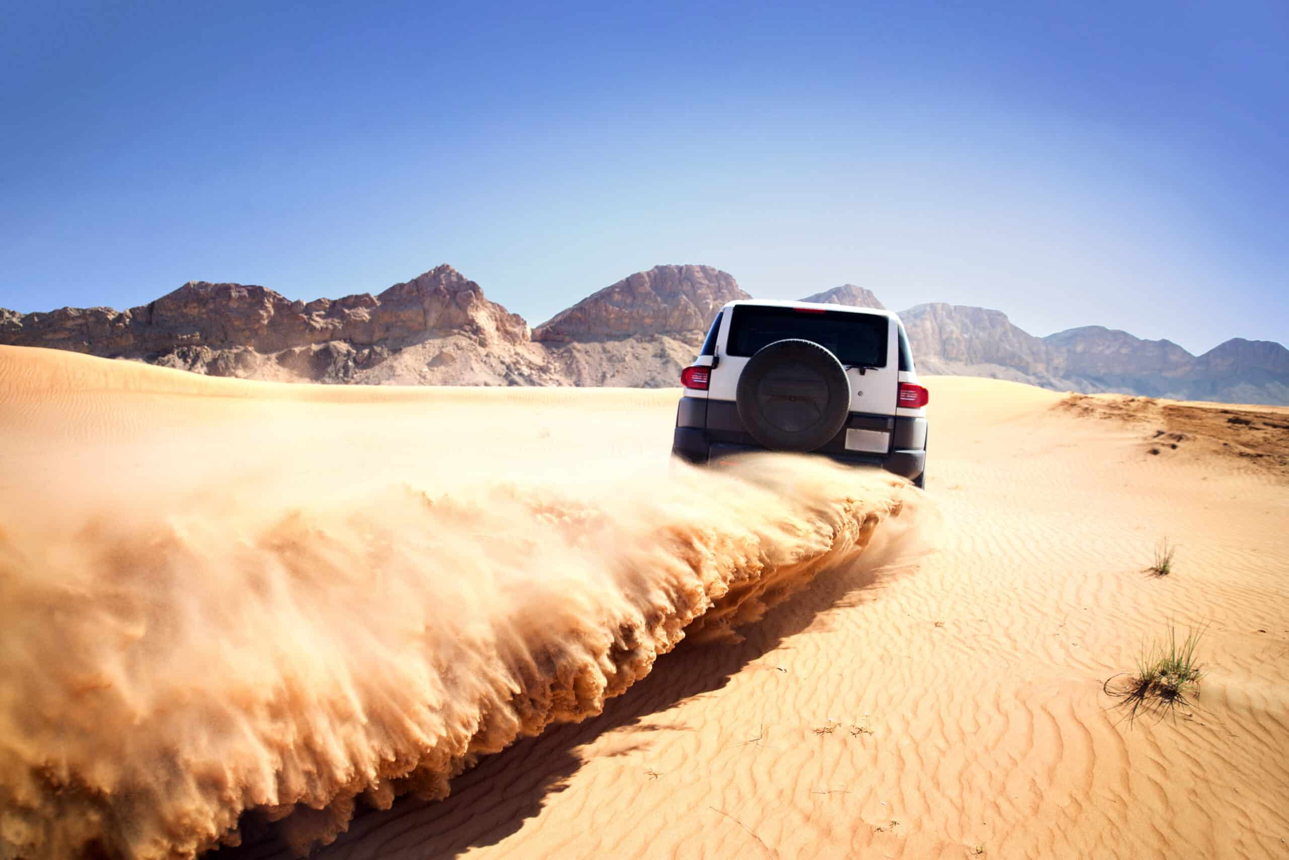 Ein weißer Geländewagen rast durch eine Sandwüste und wirbelt vor der Kulisse schroffer Berge unter einem klaren blauen Himmel eine Sandspur auf. © Fotografie Tomas Rodriguez