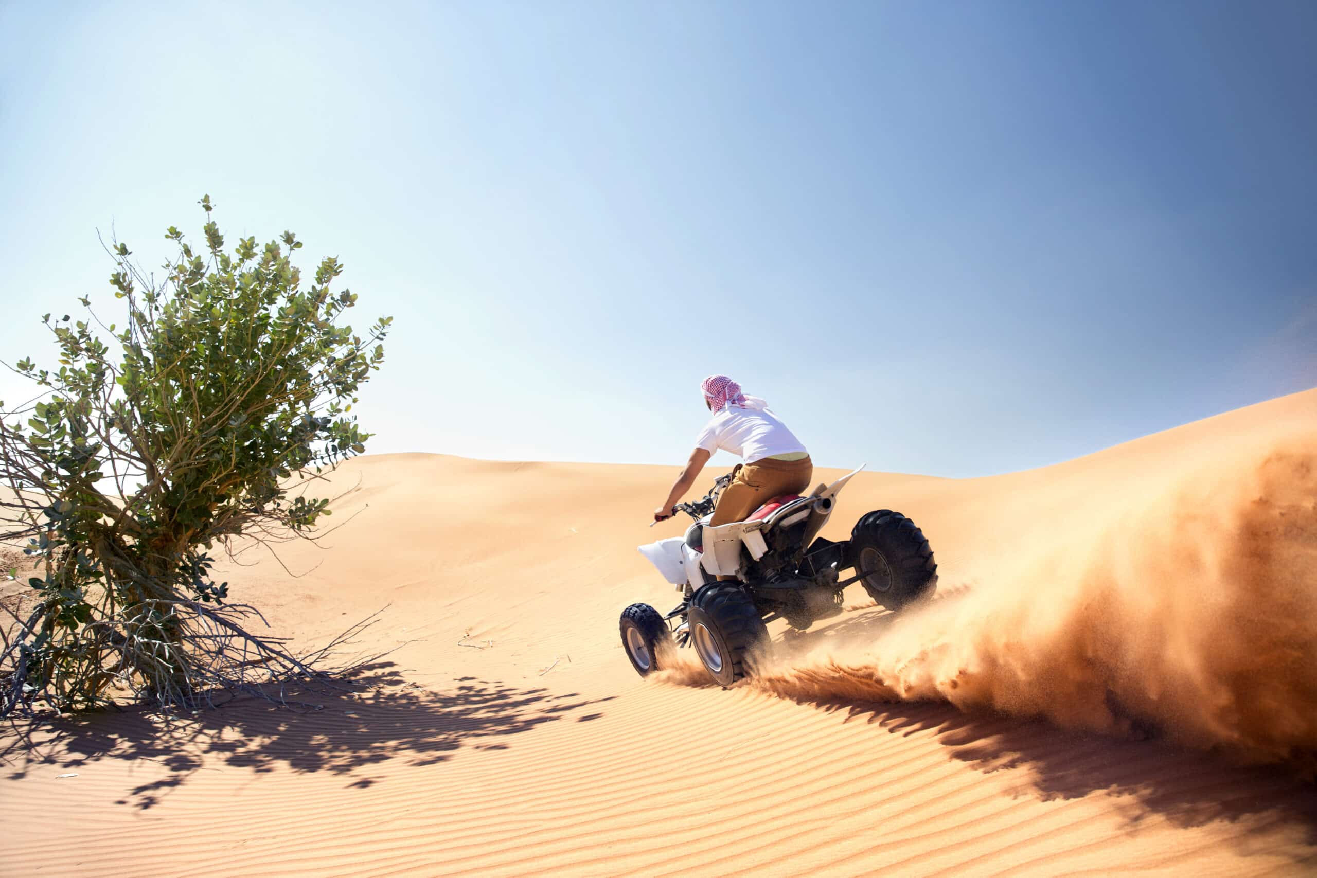 Eine Person in traditioneller Kleidung fährt auf einem Quad durch die Sanddünen der Wüste und wirbelt dabei eine Sandspur auf. Links ist unter einem klaren blauen Himmel ein grüner Busch zu sehen. © Fotografie Tomas Rodriguez