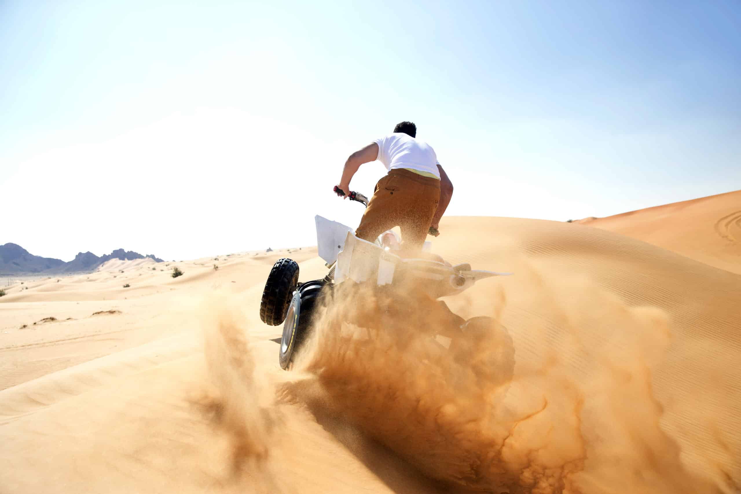 Ein Motorradfahrer fährt mit einem Dirtbike unter einem klaren blauen Himmel in einer Wüstenlandschaft eine Sanddüne hinauf und wirbelt dabei eine Sandwolke auf. © Fotografie Tomas Rodriguez