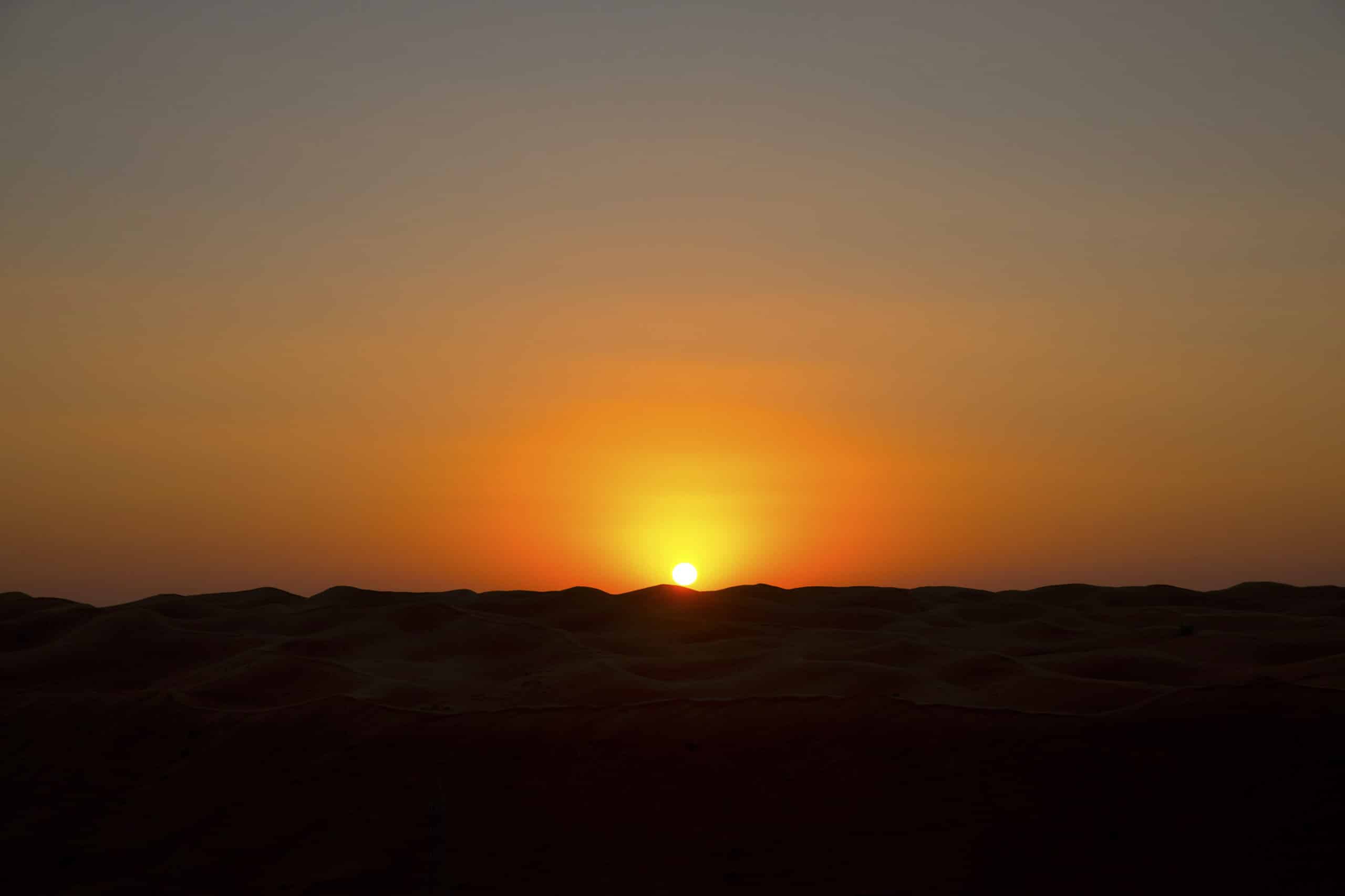 Ein ruhiger Sonnenuntergang in der Wüste, bei dem die Sonne teilweise hinter dem Horizont verschwindet und einen warmen orangefarbenen Schein über die glatten, welligen Sanddünen wirft. © Fotografie Tomas Rodriguez