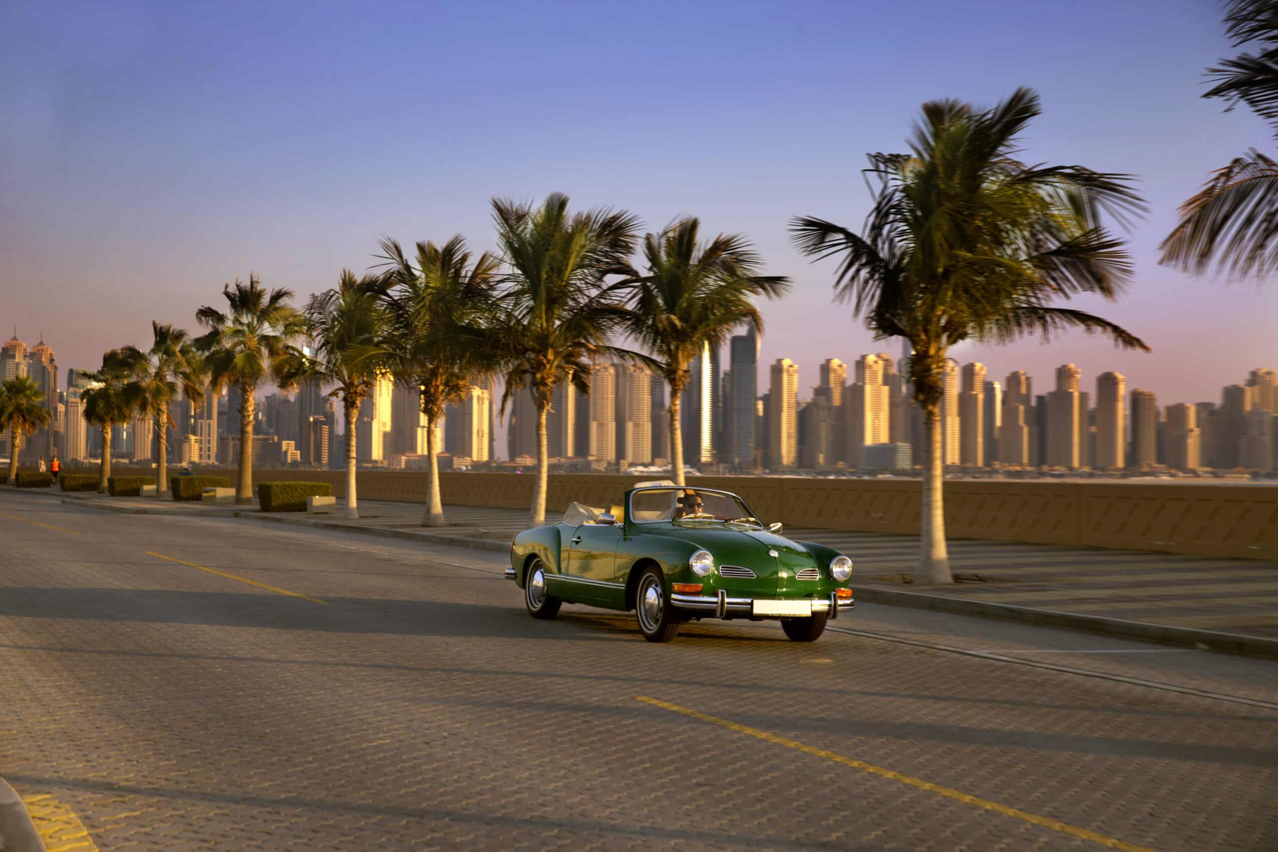 Ein grünes Oldtimer-Cabrio fährt bei Sonnenuntergang einen palmengesäumten Boulevard entlang, im Hintergrund die moderne Skyline einer Stadt. © Fotografie Tomas Rodriguez