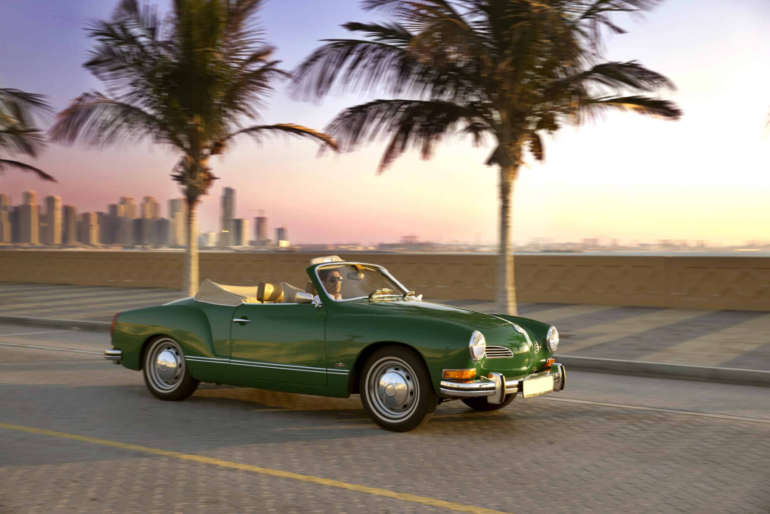 Ein grünes Oldtimer-Cabrio fährt bei Sonnenuntergang eine Küstenstraße entlang, im Hintergrund sind Palmen zu sehen und in der Ferne sieht man die Skyline einer Stadt. © Fotografie Tomas Rodriguez