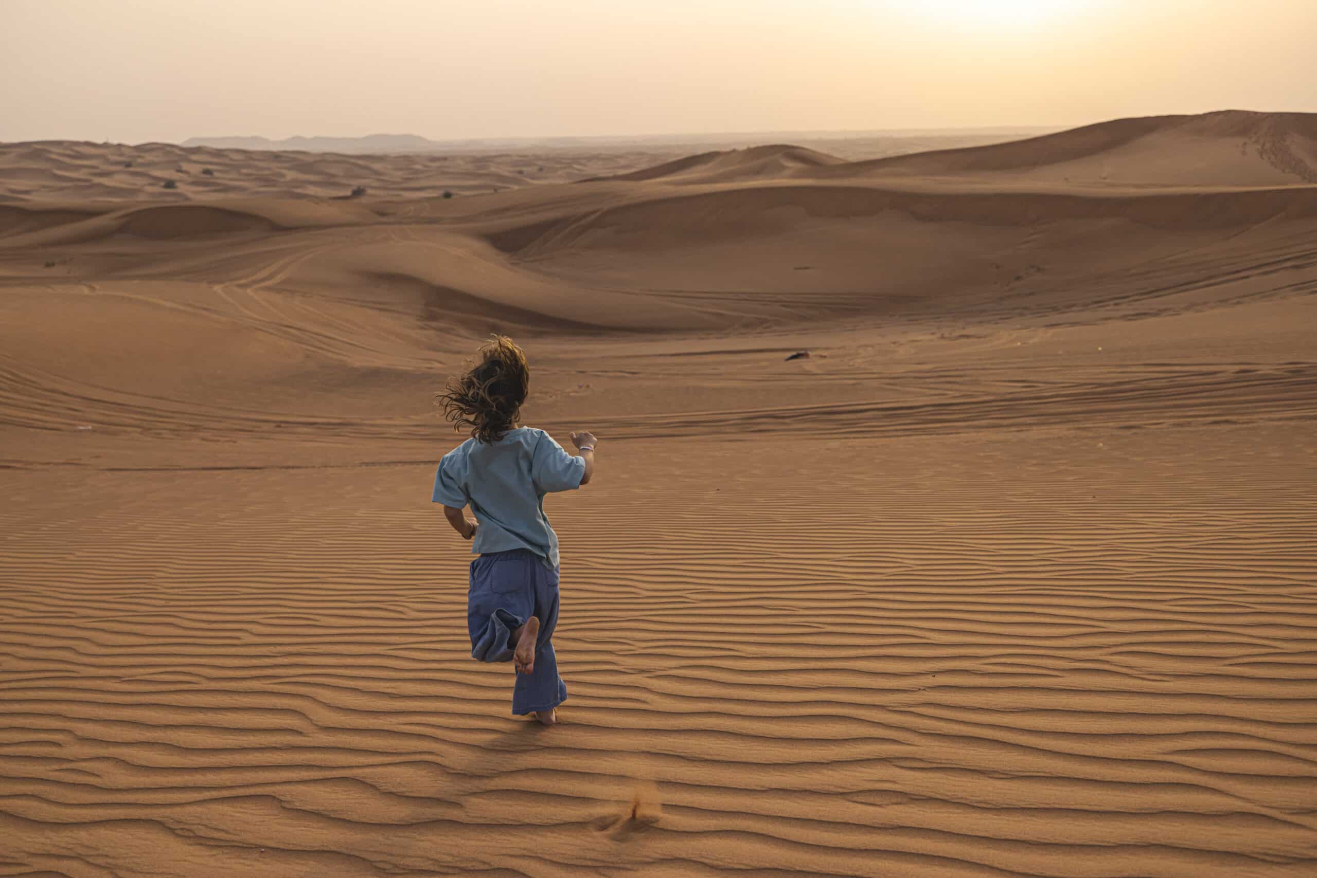 Ein kleines Kind läuft bei Sonnenuntergang über Sanddünen und fängt die Landschaft mit einem Smartphone ein, während seine Haare im Wind wehen. Die Wüste strahlt in einem warmen orangefarbenen Glanz. © Fotografie Tomas Rodriguez