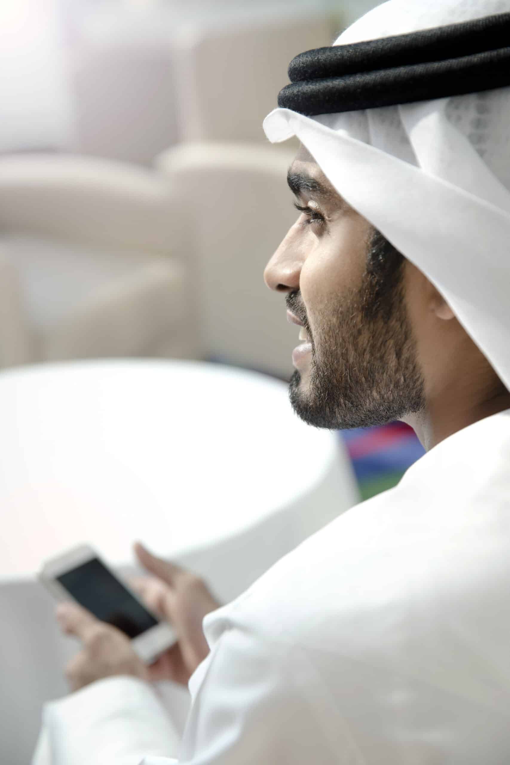 Ein Mann in traditioneller emiratischer Kleidung, eine weiße Kandura und einen schwarzen Agal tragend, hält ein Smartphone und blickt nachdenklich in die Ferne. © Fotografie Tomas Rodriguez