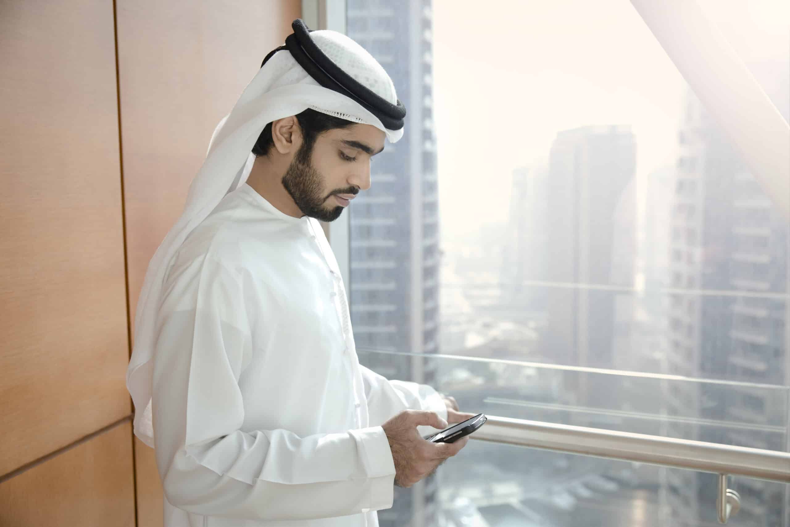 Ein Mann in traditioneller emiratischer Kleidung steht an einem Fenster mit Blick auf die Stadtlandschaft und konzentriert sich auf sein Smartphone. © Fotografie Tomas Rodriguez
