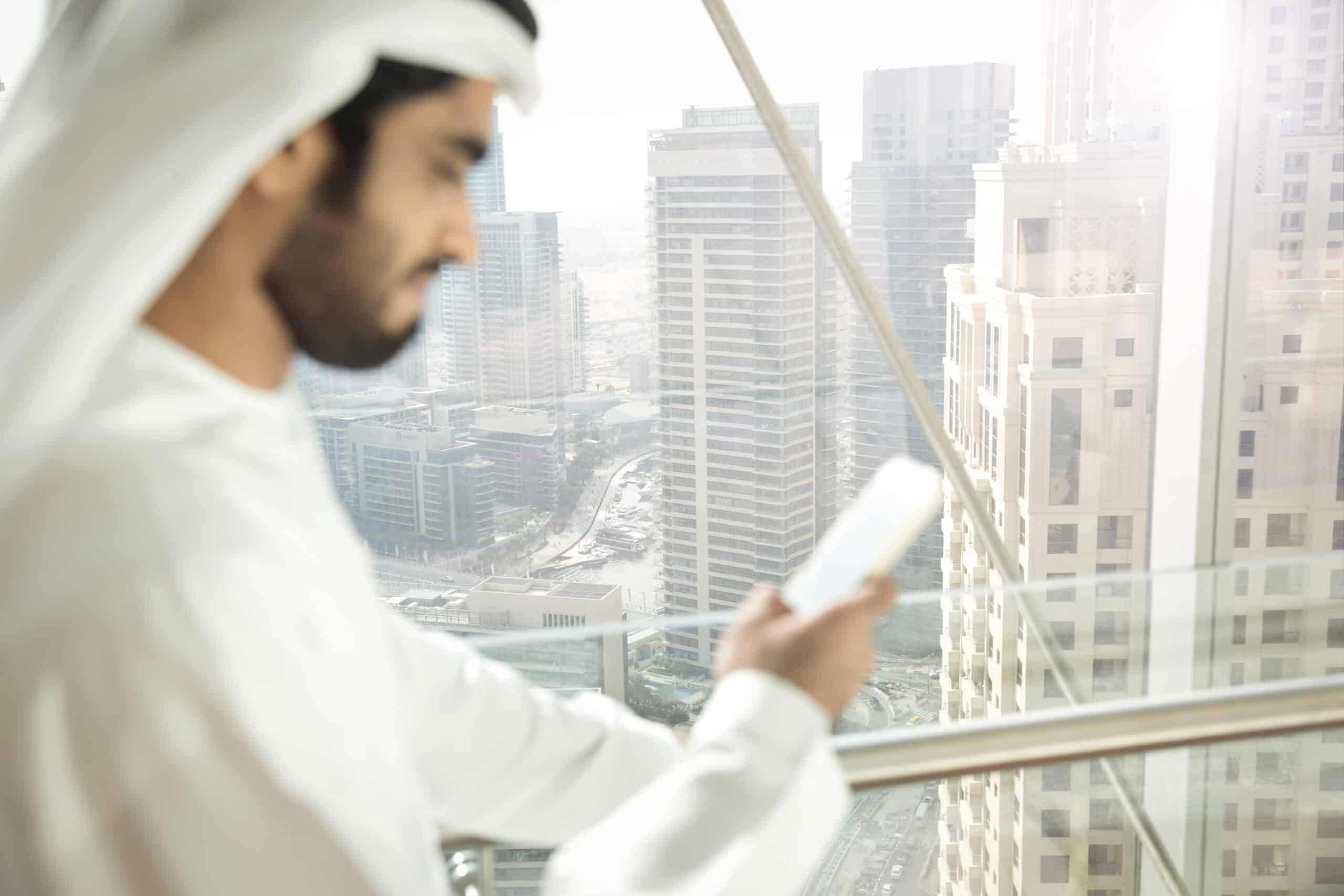 Ein Mann in traditioneller emiratischer Kleidung schaut auf sein Smartphone, während er an einem großen Fenster steht und auf eine moderne Stadtlandschaft mit Wolkenkratzern blickt. © Fotografie Tomas Rodriguez