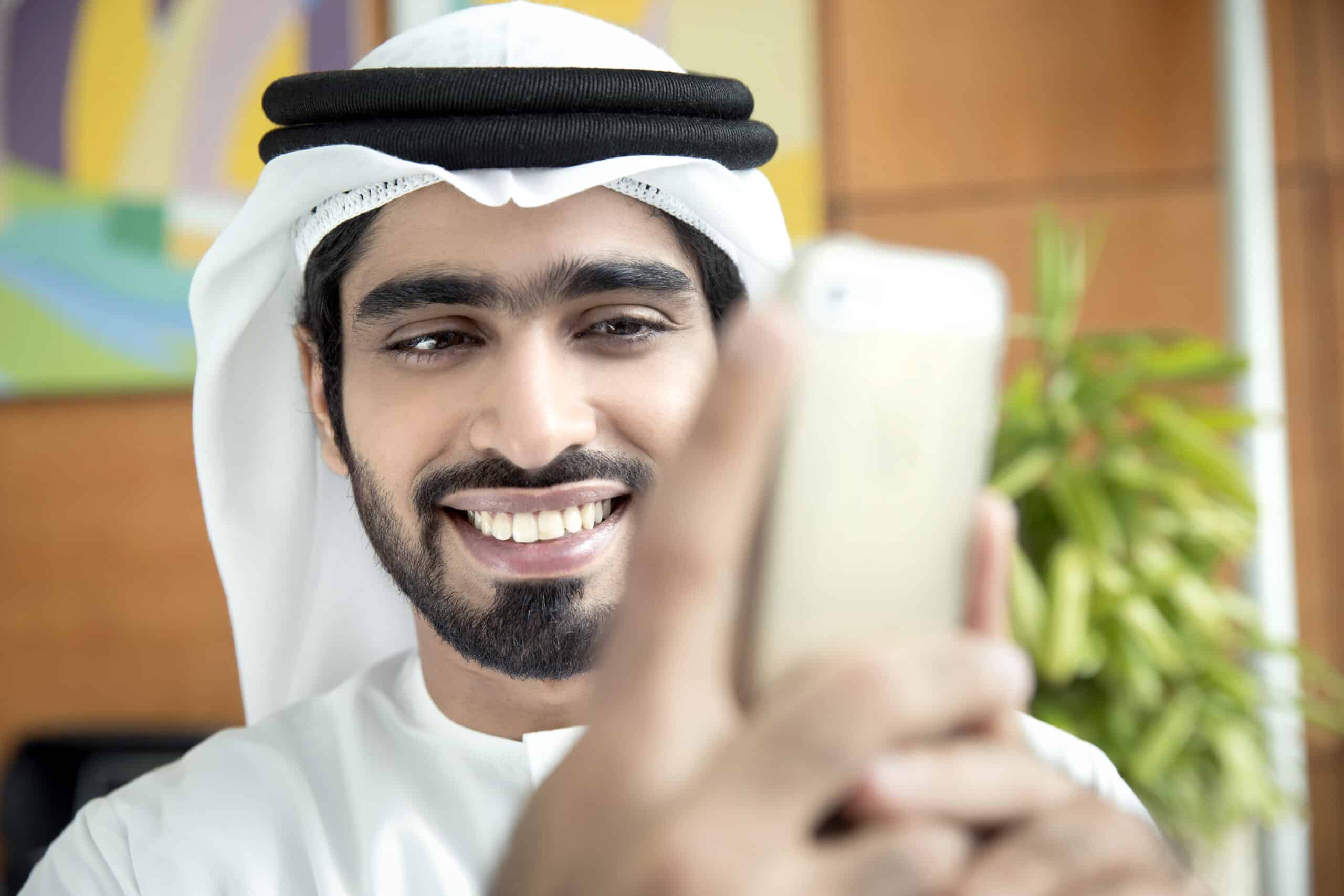 Ein lächelnder Mann in traditioneller emiratischer Kleidung hält ein Smartphone, scheint ein Selfie zu machen oder mit dem Telefon zu interagieren. Er ist in einem Gebäude mit farbenfrohen Kunstwerken im Hintergrund. © Fotografie Tomas Rodriguez