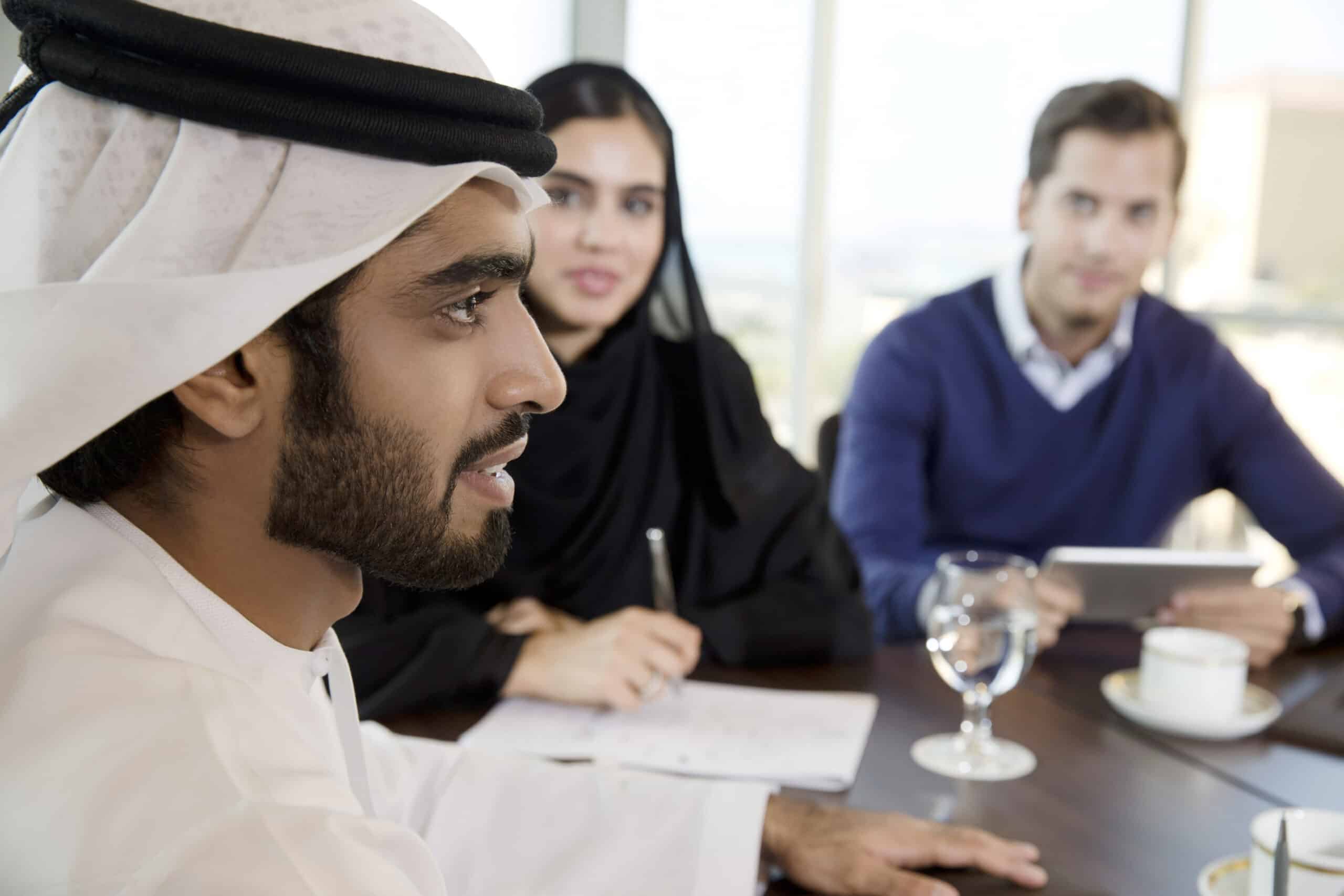 Ein Mann in traditioneller emiratischer Kleidung und eine Frau im schwarzen Hijab sitzen mit einem weißen Mann im blauen Pullover an einem Besprechungstisch in einem hellen, modernen Büroumfeld. © Fotografie Tomas Rodriguez