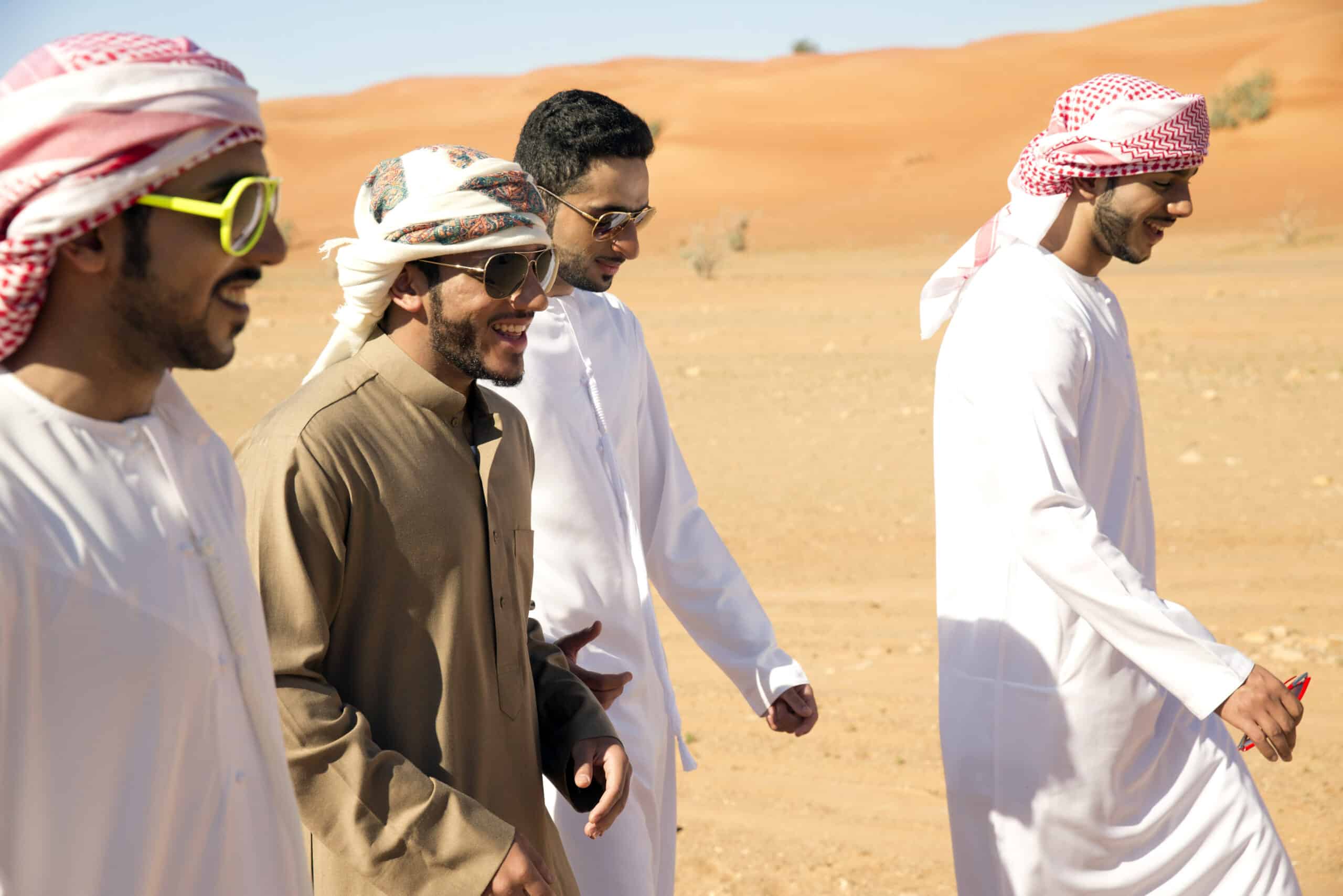Vier arabische Männer in traditionellen weißen Gewändern und karierten Kopftüchern gehen und unterhalten sich gemeinsam in einer Wüstenlandschaft, einer davon trägt eine modische Sonnenbrille. © Fotografie Tomas Rodriguez