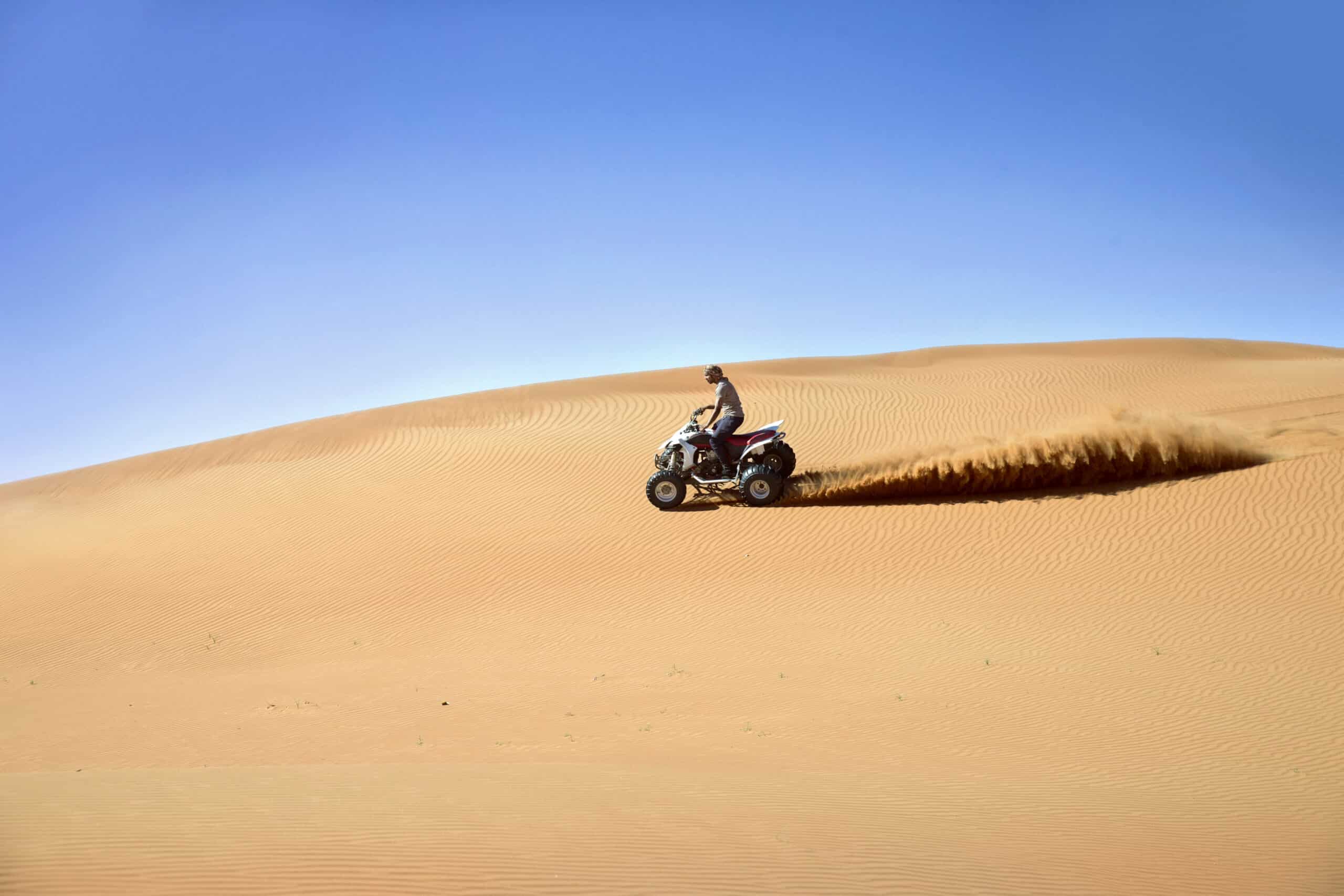 Eine Person fährt mit einem Geländefahrzeug über eine Sanddüne in einer riesigen Wüste und hinterlässt unter einem klaren blauen Himmel eine Staubspur. © Fotografie Tomas Rodriguez