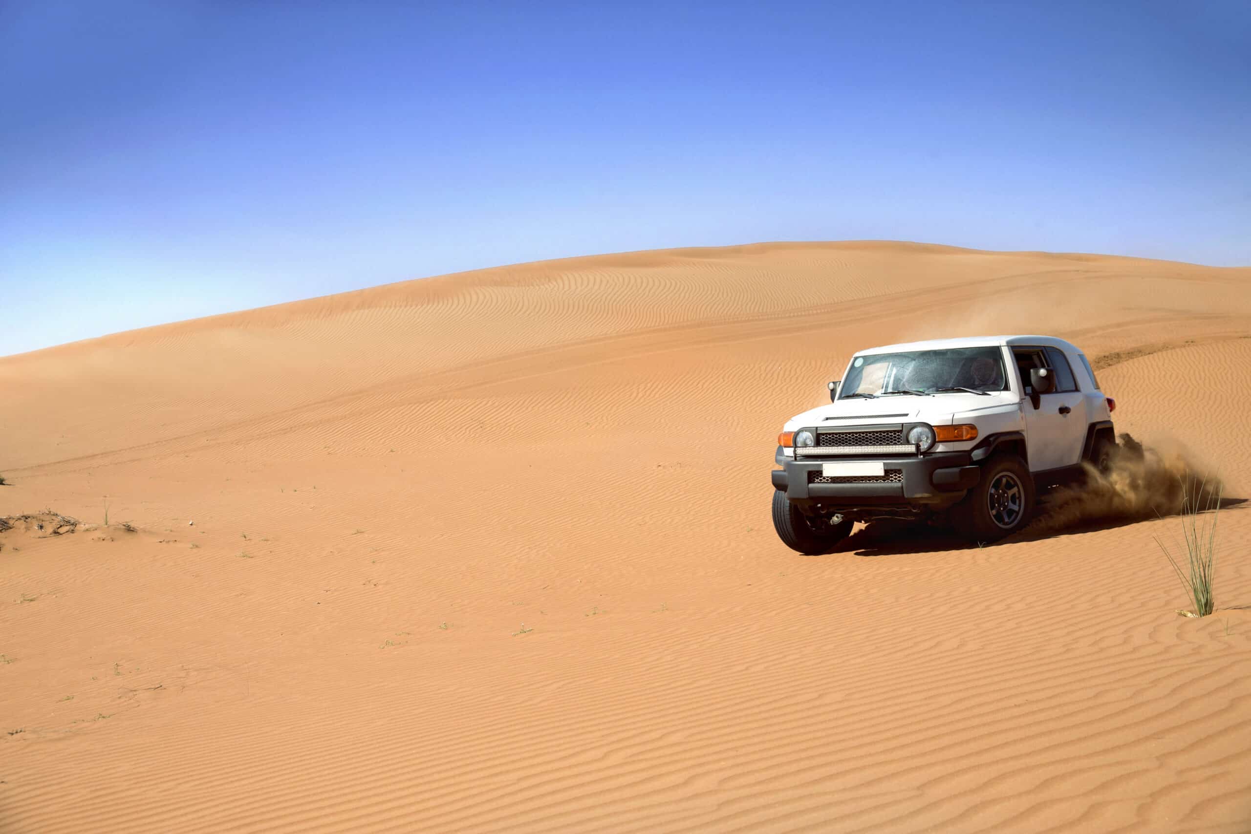 Ein weißer SUV fährt unter einem klaren blauen Himmel durch eine sandige Wüstenlandschaft und wirbelt dabei Sand auf. © Fotografie Tomas Rodriguez