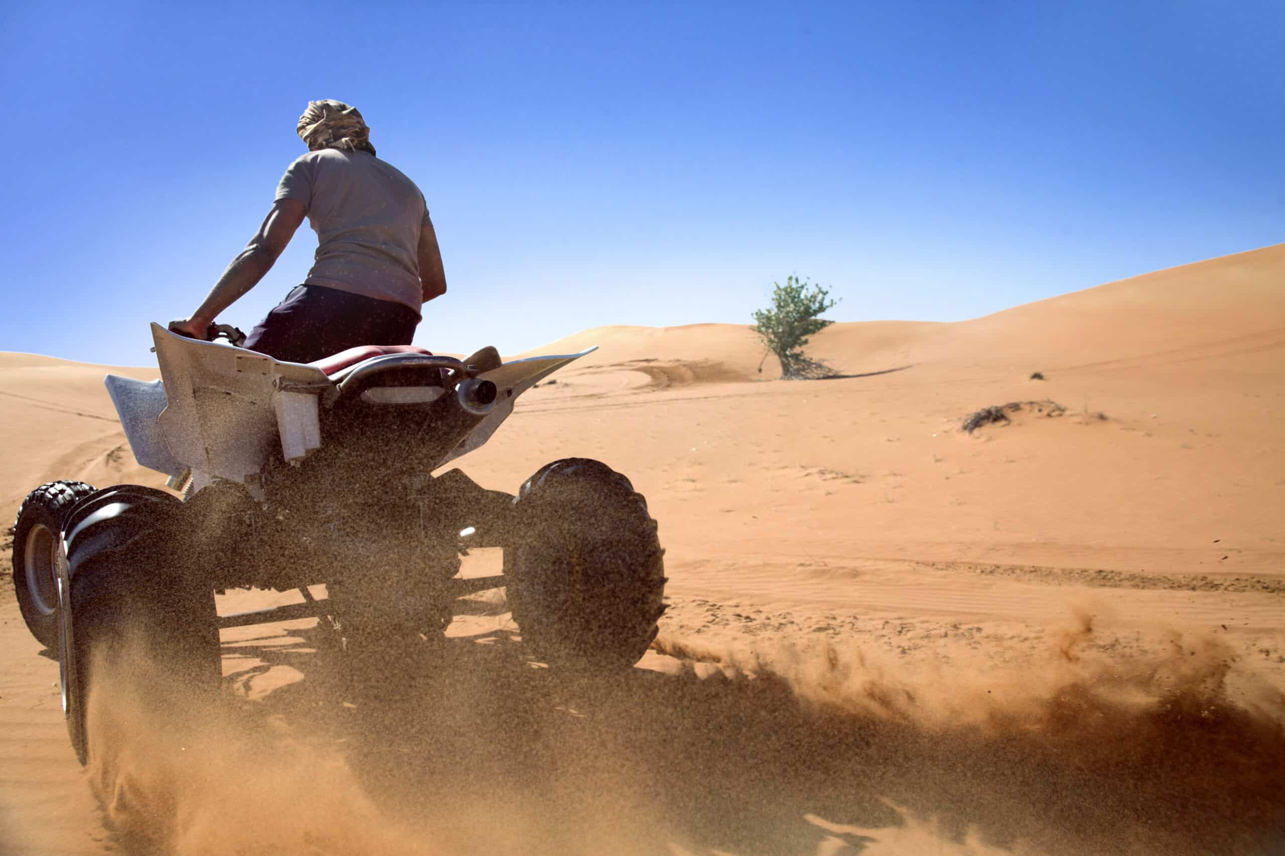 Eine Person mit weißem Hemd und Kopftuch fährt unter einem klaren blauen Himmel auf einem Quad über Sanddünen und wirbelt dabei Sandspuren auf. © Fotografie Tomas Rodriguez