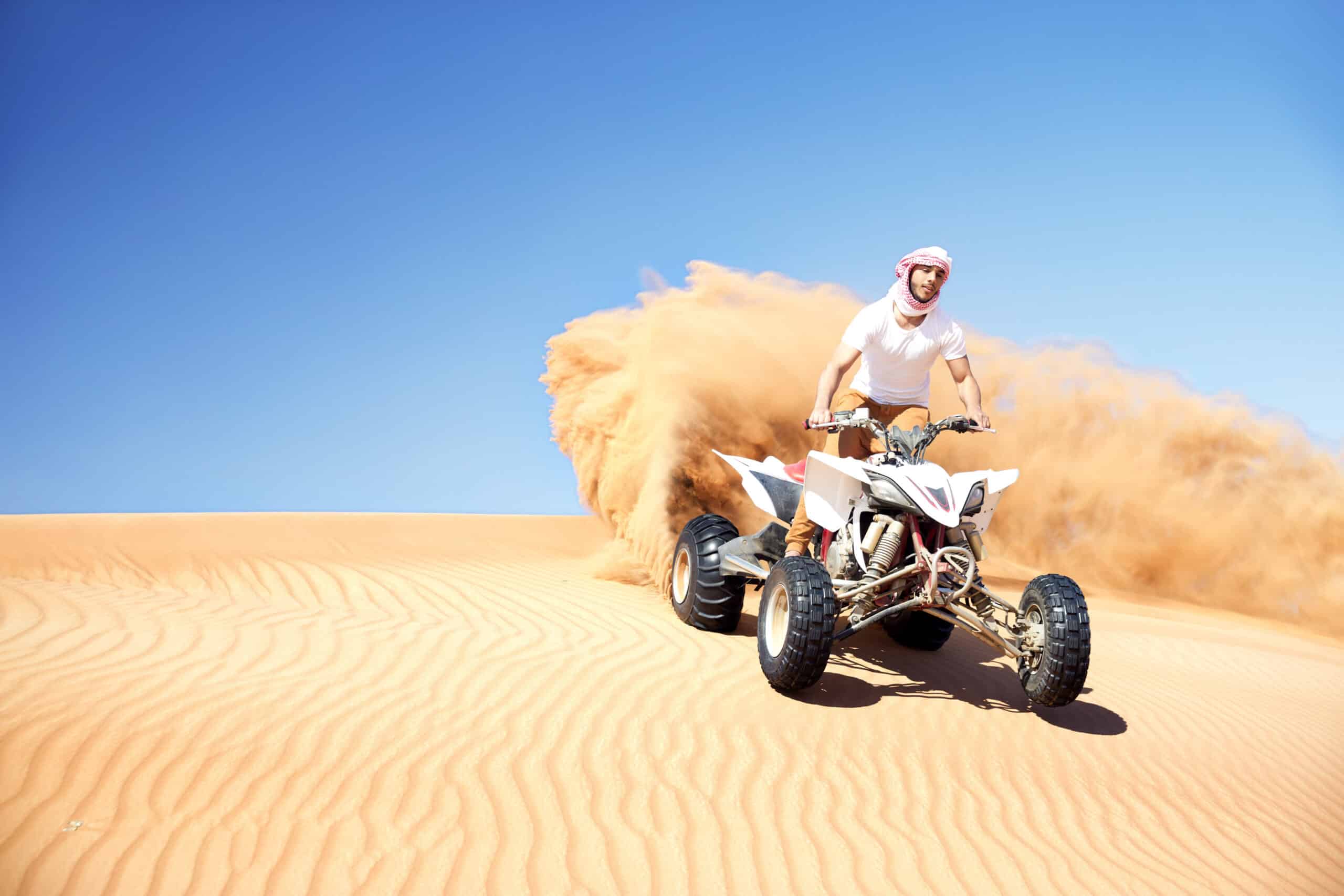 Eine Person in traditioneller nahöstlicher Kleidung fährt unter einem klaren blauen Himmel auf einem Quad durch Sanddünen und wirbelt dabei eine Sandspur auf. © Fotografie Tomas Rodriguez